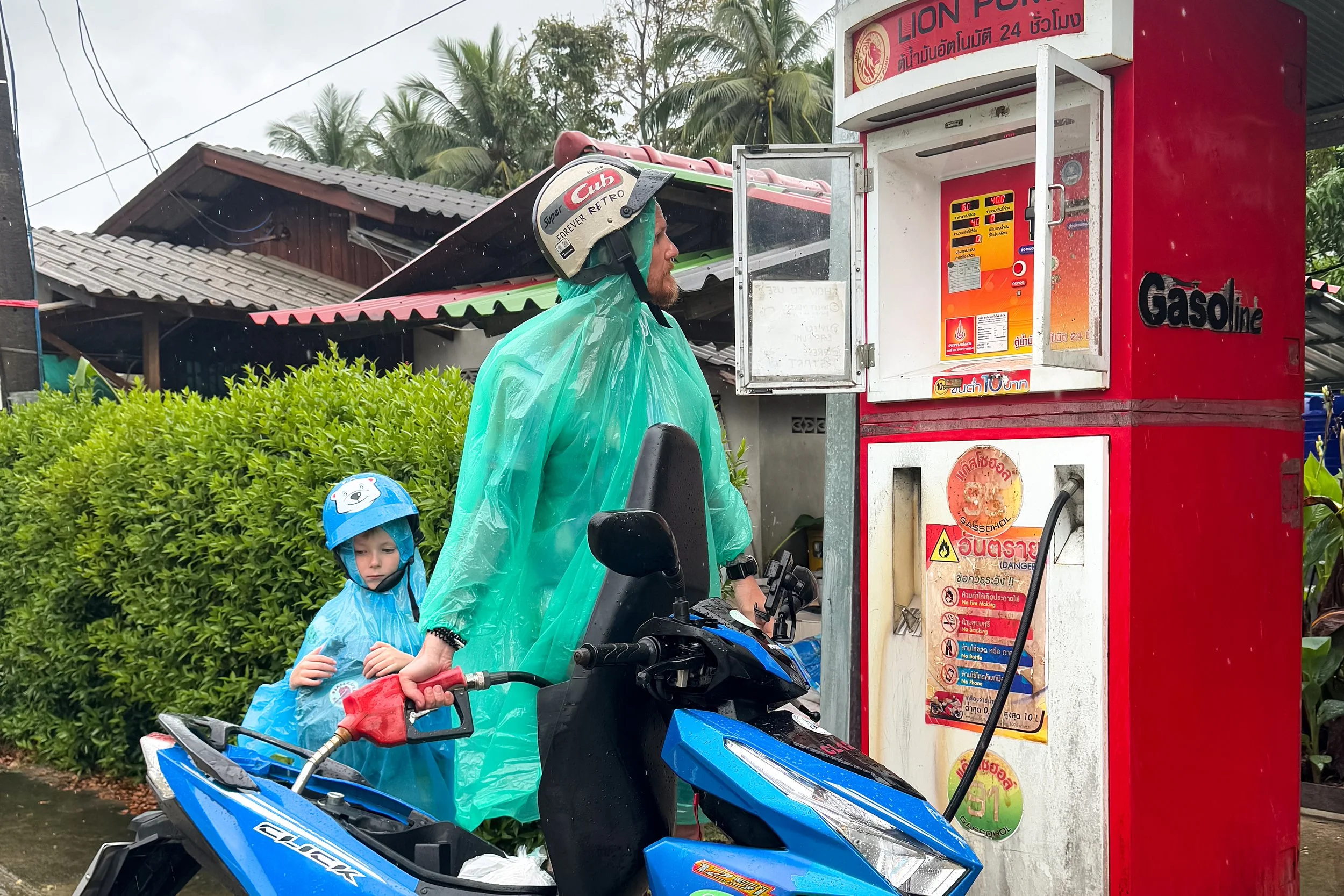 A man and a child on a blue motorcycle at a gas station, refueling. The man is wearing a white helmet, green rain poncho, and holding the motorcycle handlebar, while the child is in a blue raincoat with a hood featuring a cartoon character. A red fuel pump with Thai text and symbols is nearby, with tropical greenery and houses in the background.