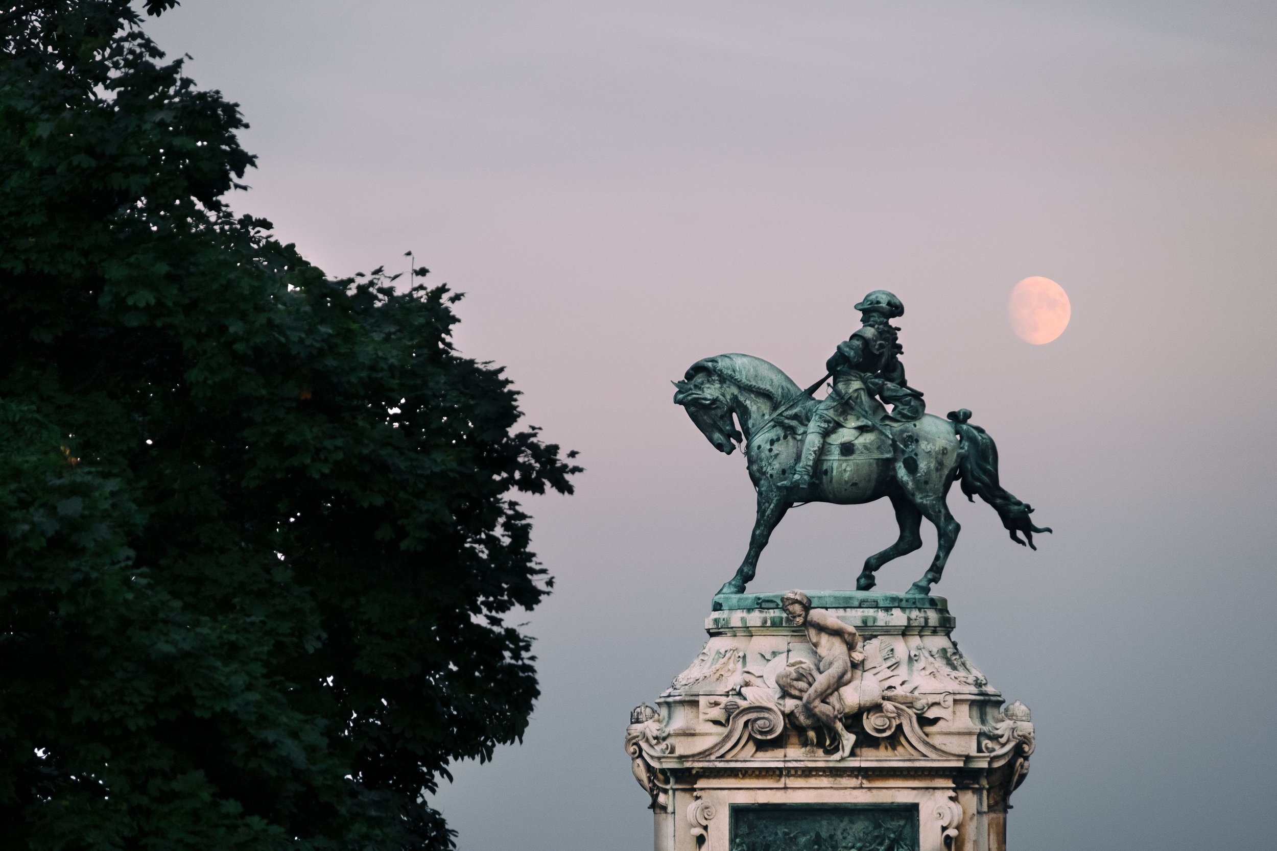 A bronze statue of a man on a horse on a decorative pedestal, with a large, bright moon in the cloudy sky behind it, and a dark leafy tree on the left.