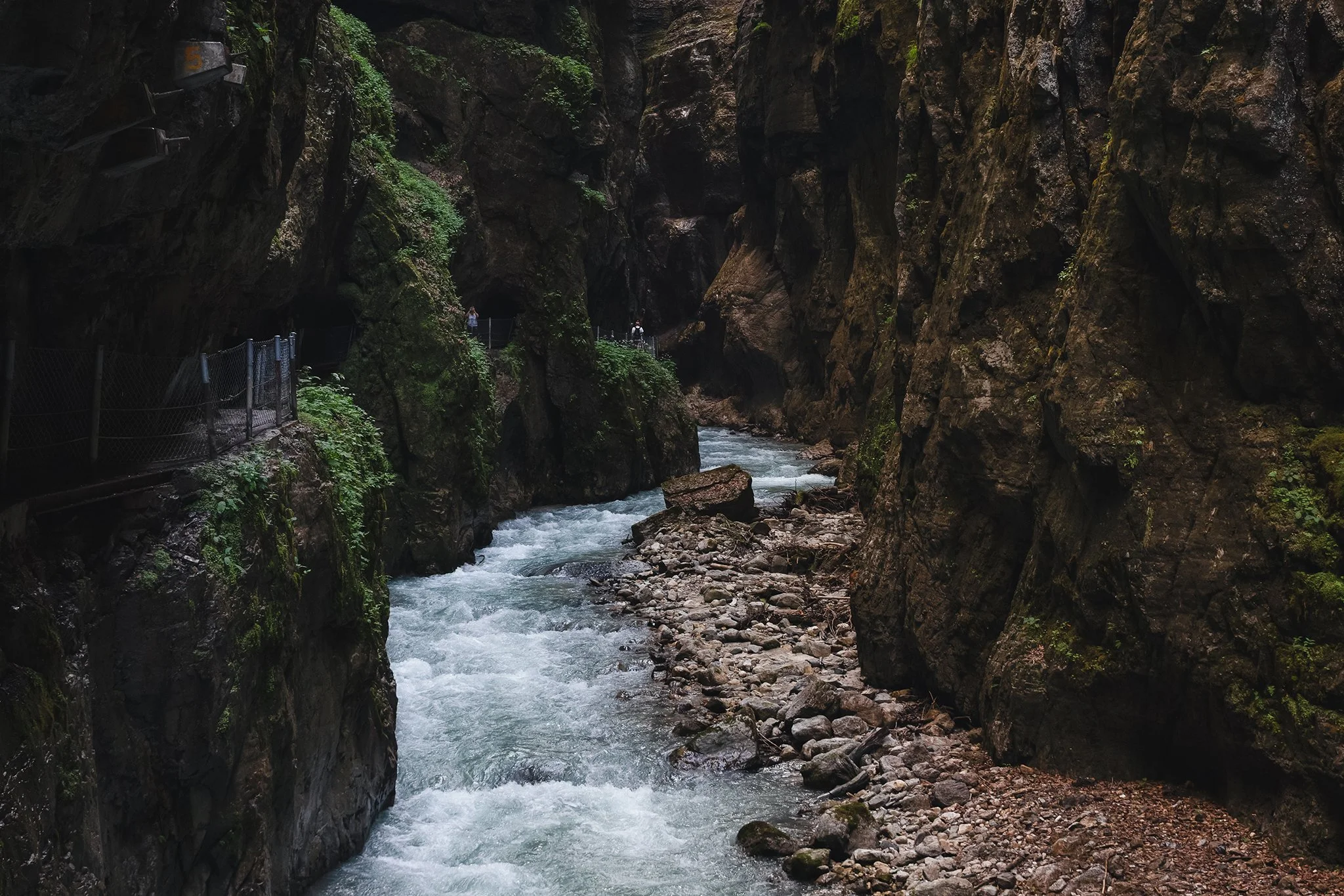 A narrow canyon with steep, dark rocky walls covered in patches of green moss and vegetation. A fast-moving river flows through the canyon at the bottom, surrounded by rocks and boulders. There are a few visitors walking along a walkway with a fence on the left side of the image.