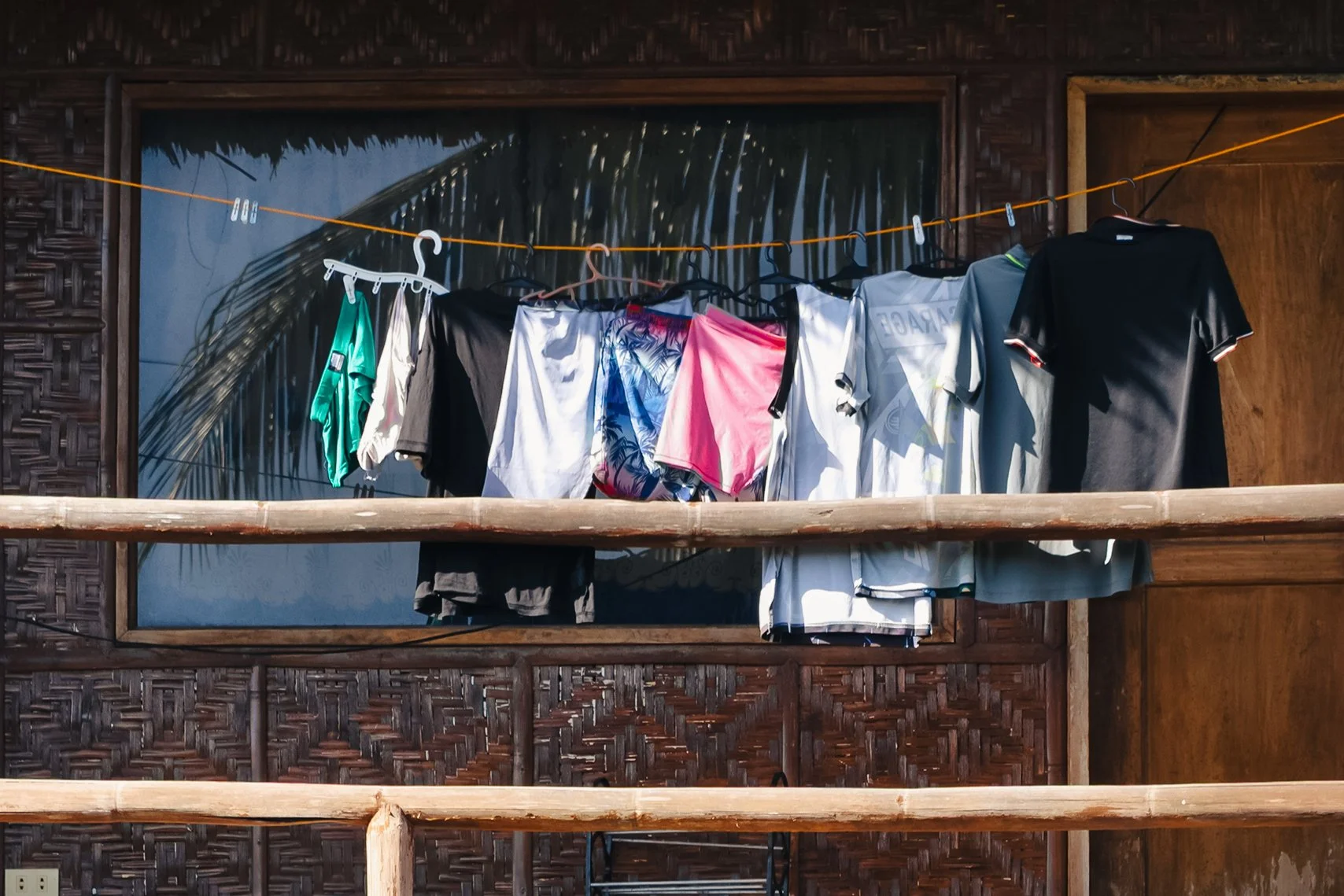 Clothes hanging on a clothesline in front of a wooden wall with a framed tropical scene, with a wooden balcony railing in the foreground.