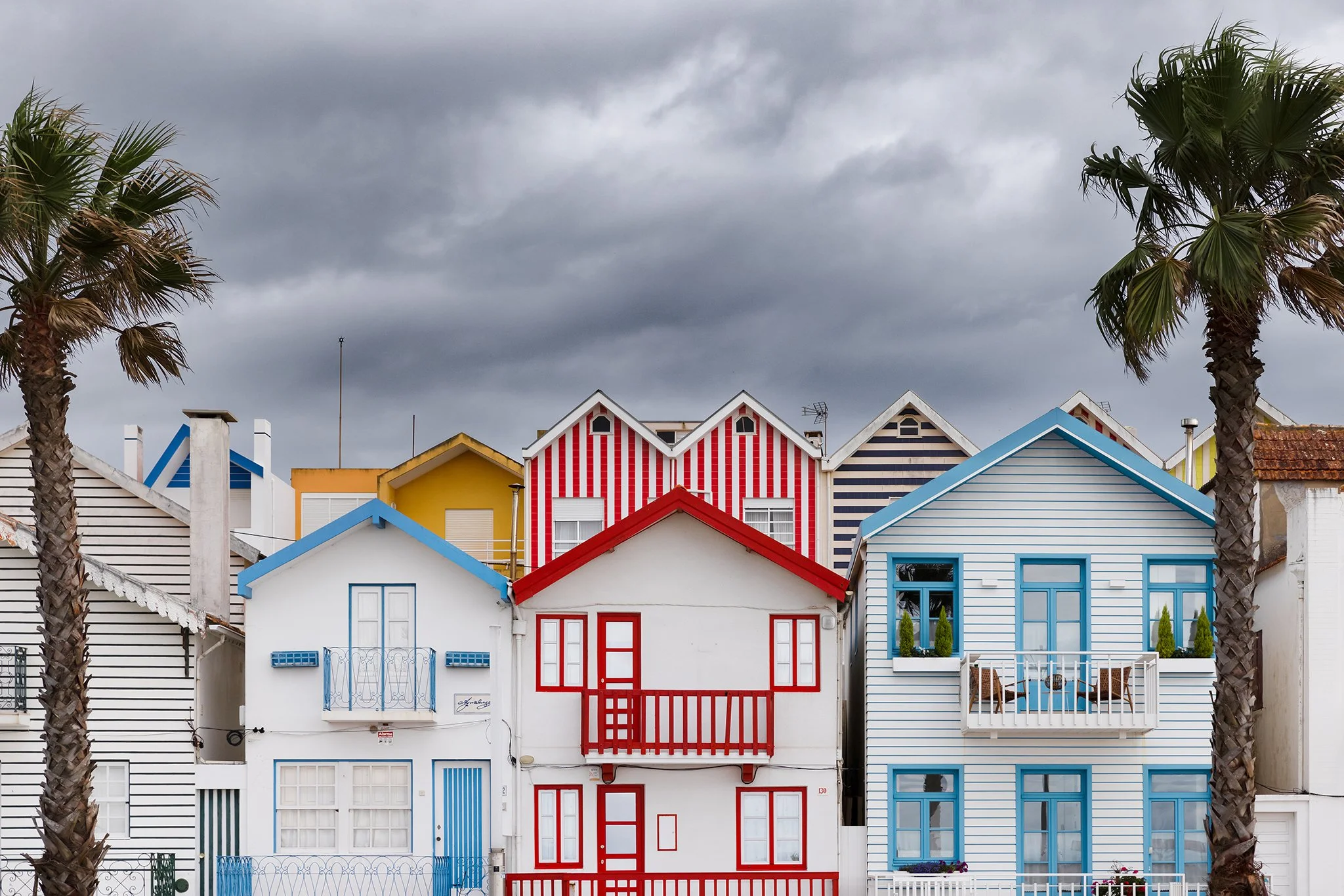 Colorful row houses with steep roofs and palm trees under a cloudy sky.