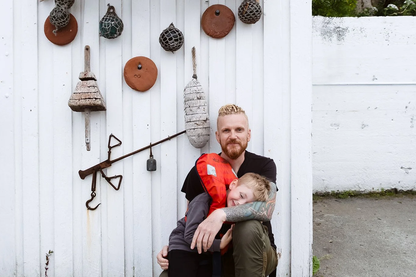 A man with a beard hugging a young boy with blond hair in front of a white wooden wall decorated with rustic farm tools and farm-related objects.
