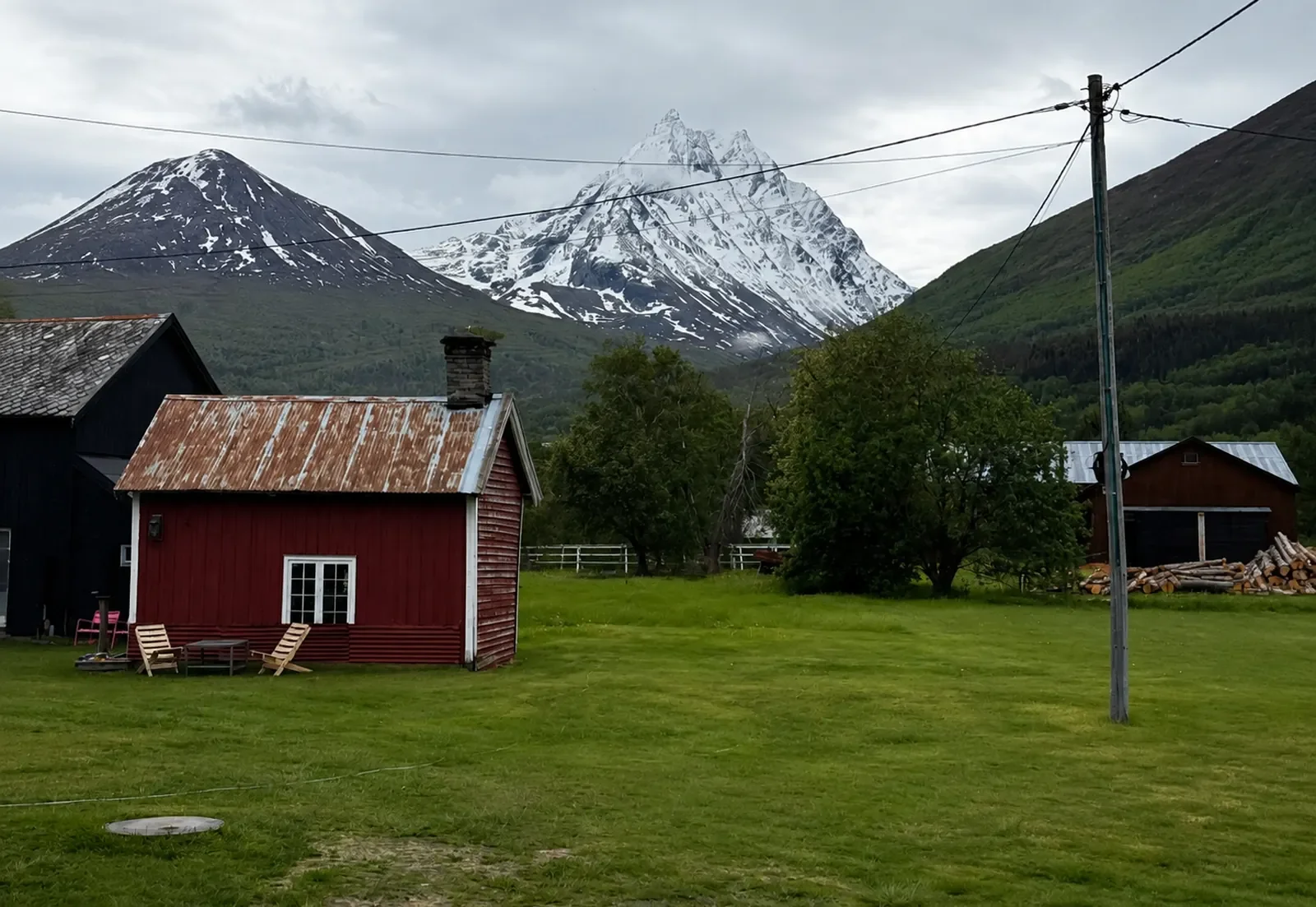 A rural scene with a red wooden house, green grass, trees, a utility pole, and a mountain range with snow-capped peaks in the background under a cloudy sky.