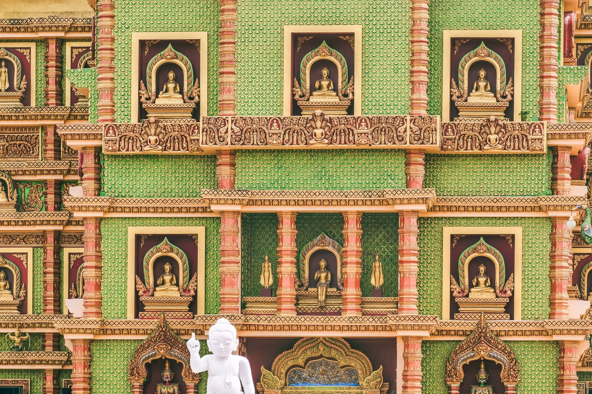 Colorful exterior of a Thai temple with multiple Buddha statues and intricate decorations, including a large white Buddha statue in the foreground and a vibrant green patterned background.