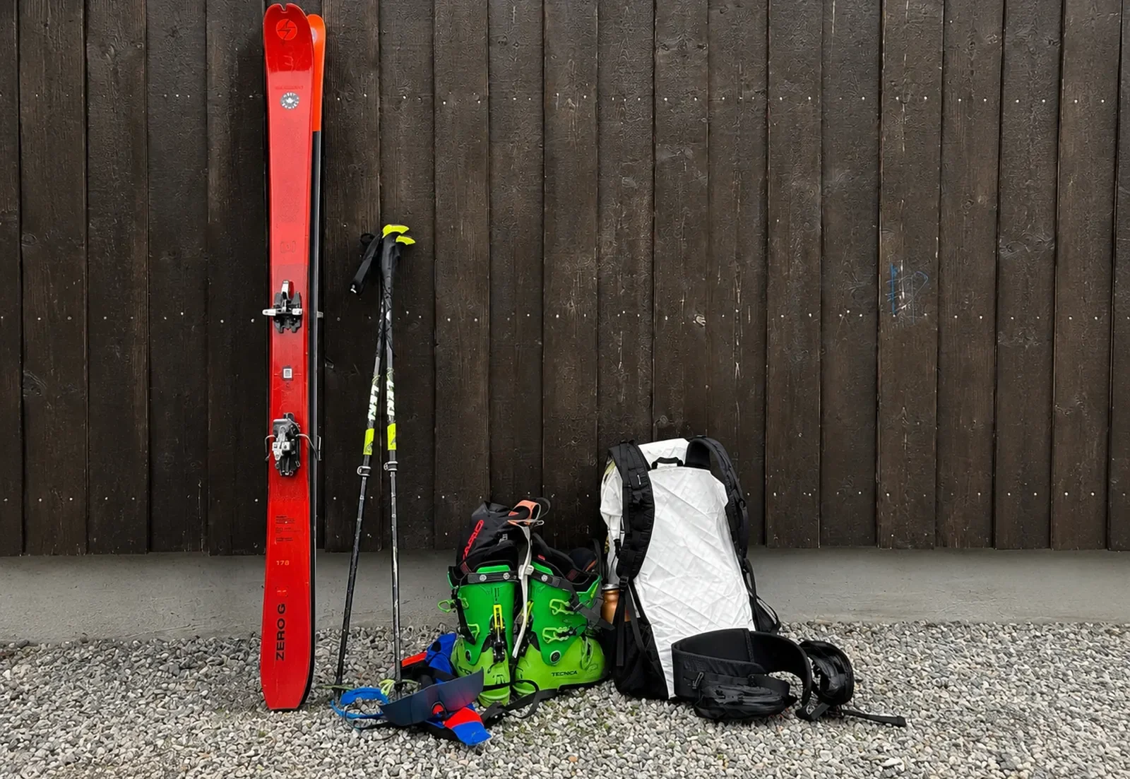 Ski equipment and gear including a pair of green ski boots, a white backpack, a pair of ski poles, and a pair of skis leaning against a dark wooden fence, with small gravel on the ground.