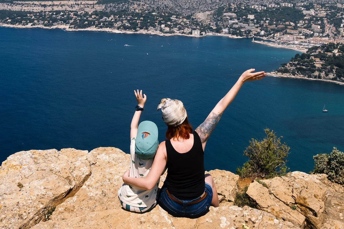 A woman and a child sitting on a rocky cliff edge overlooking a coastal city and ocean, with the woman raising her arm as if pointing toward the water.