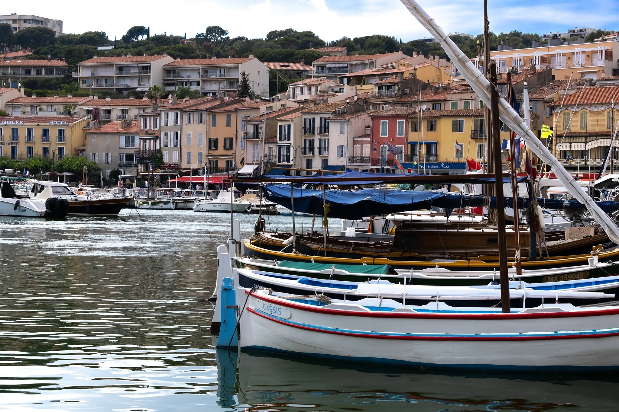 A marina with boats docked in the water and a hillside with colorful buildings in the background.