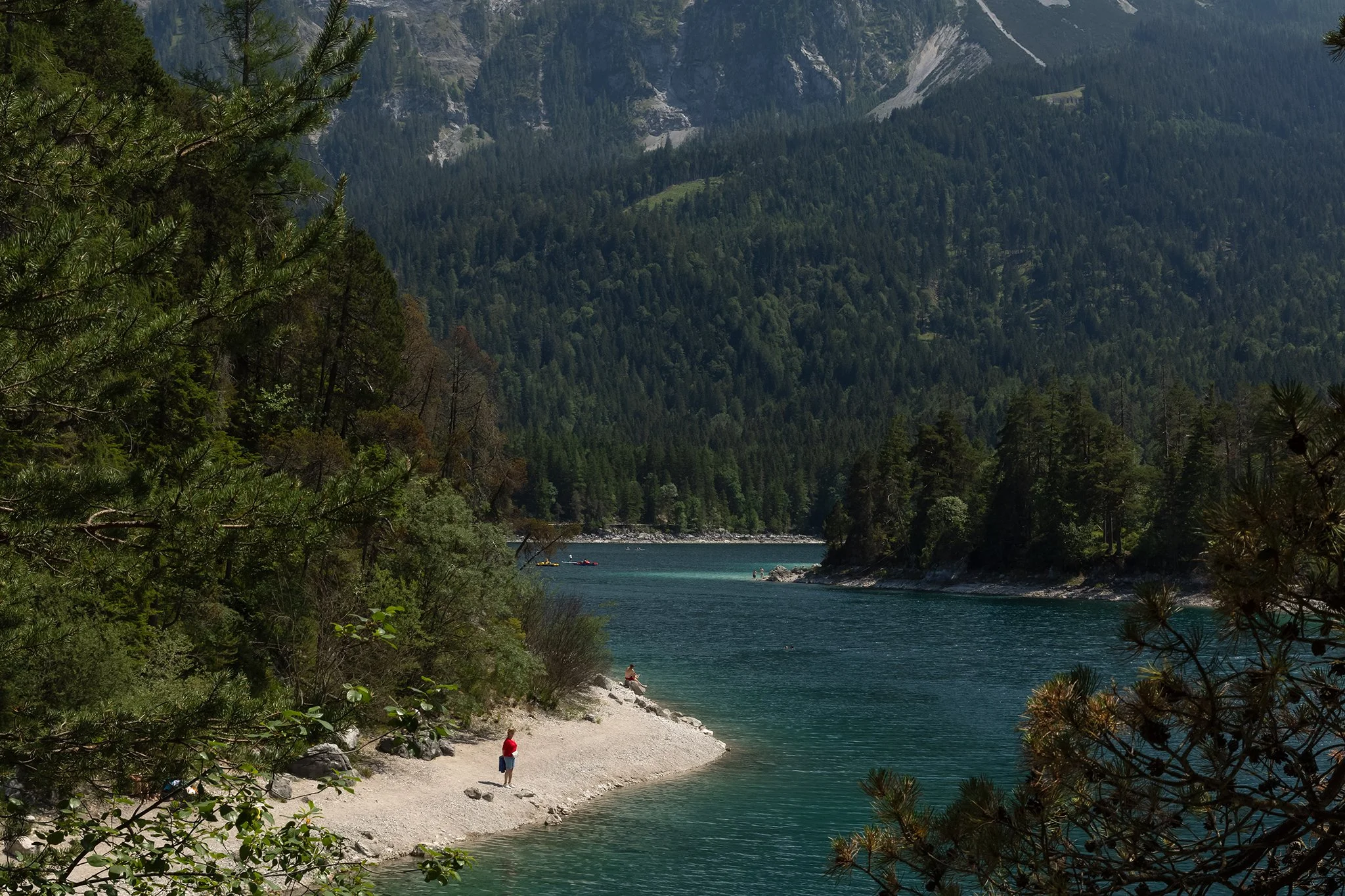 A scenic view of a lake surrounded by dense forested mountains with a small sandy beach and a few people enjoying the water and snacks, framed by pine branches.