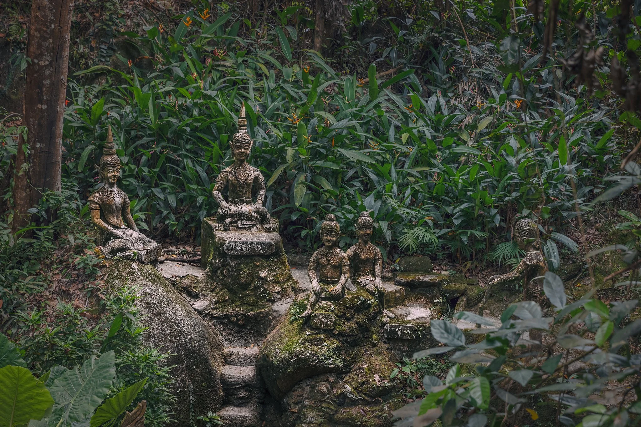Ancient stone statues of traditional Balinese figures placed along a natural outdoor stone staircase, surrounded by dense green tropical plants and foliage.