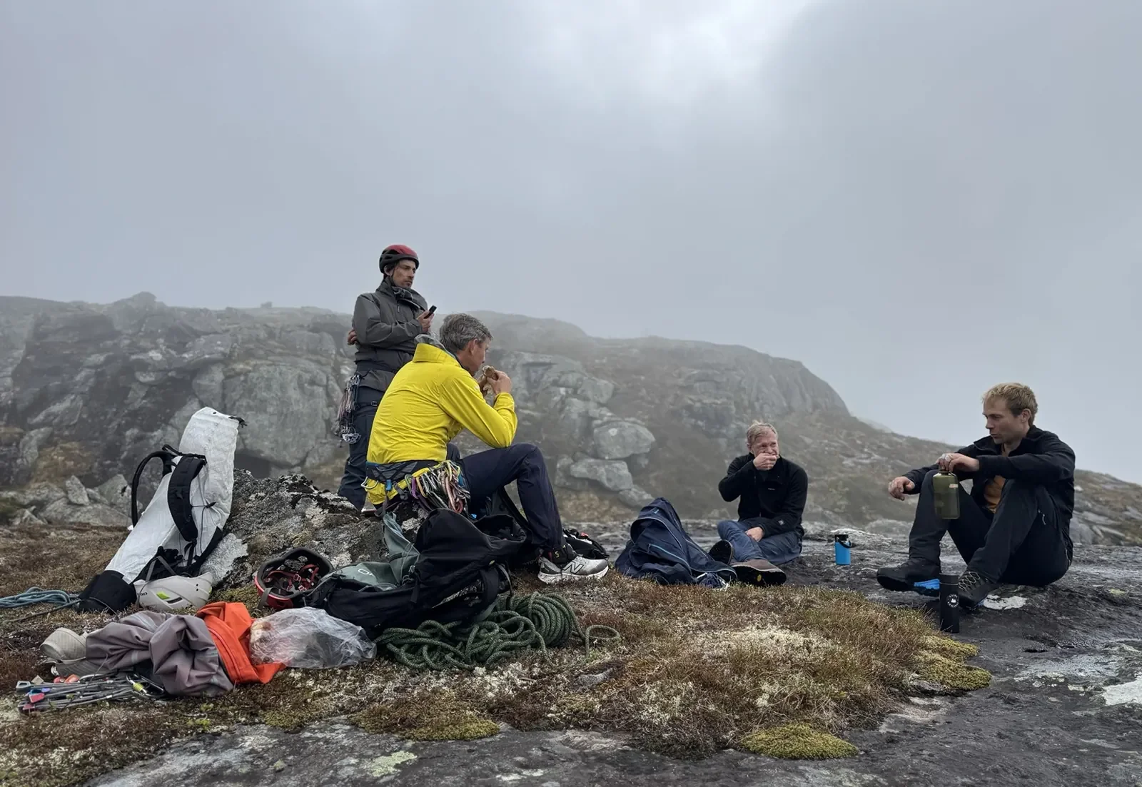 A group of five hikers resting on a rocky mountain slope with gear around them, some sitting and one standing and using a phone, in cloudy weather.