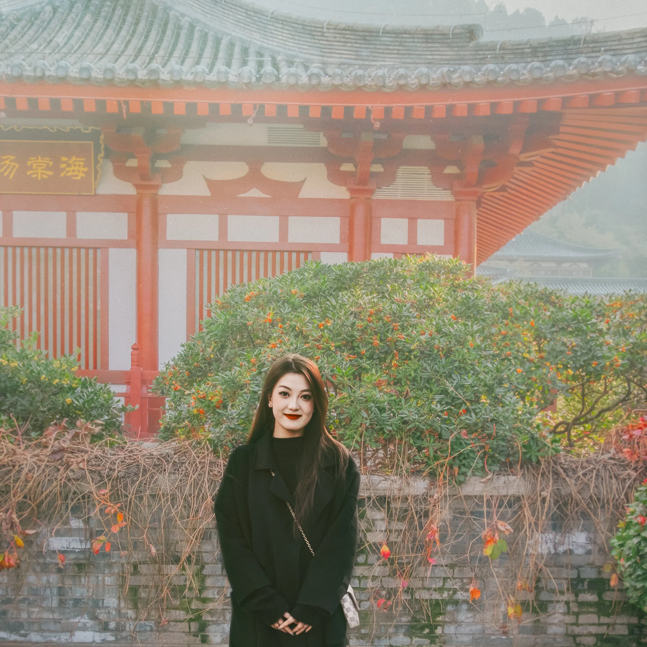 A woman in a black coat standing in front of a brick wall covered with green and red foliage, with a traditional Asian-style roof in the background.