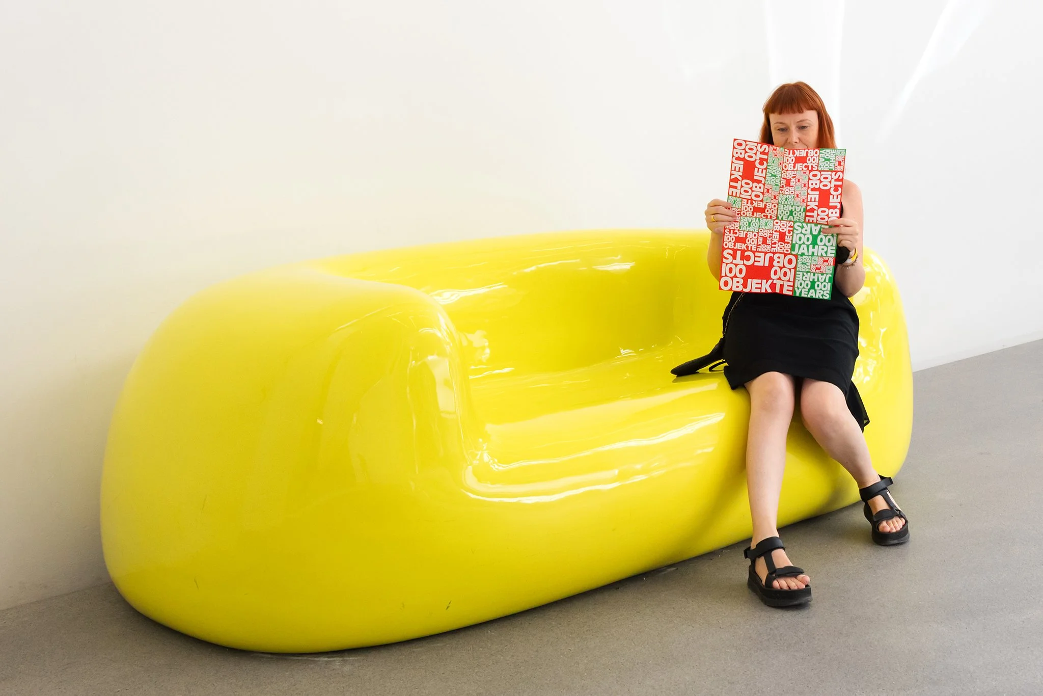 A woman with red hair wearing black clothing and sandals sitting on a large, shiny yellow sculpture with a smooth, rounded shape in an indoor art gallery, holding a red and white booklet.