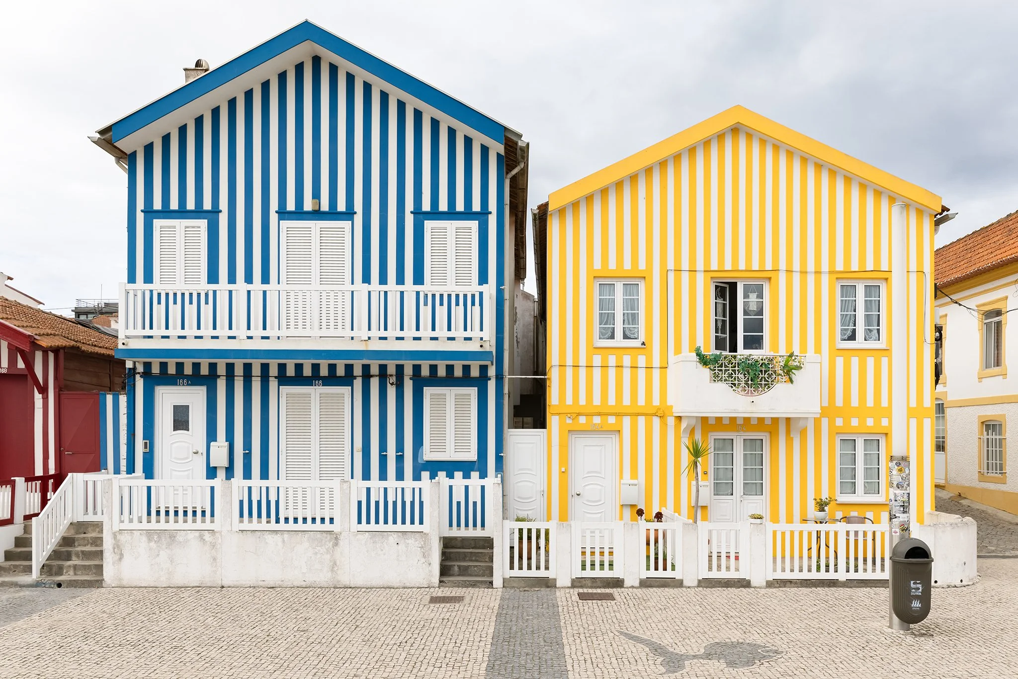 Two colorful houses with vertical stripes, one painted blue and white, the other yellow and white, with white fences in front. The houses are set against a cloudy sky.