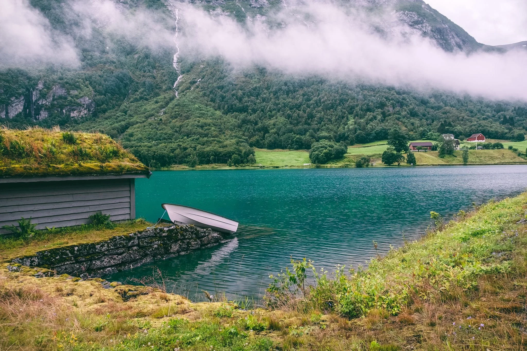 View of a mountainous landscape with fog, a lake with clear water, and a small building near the water's edge, with a boat resting on the shore in the foreground.
