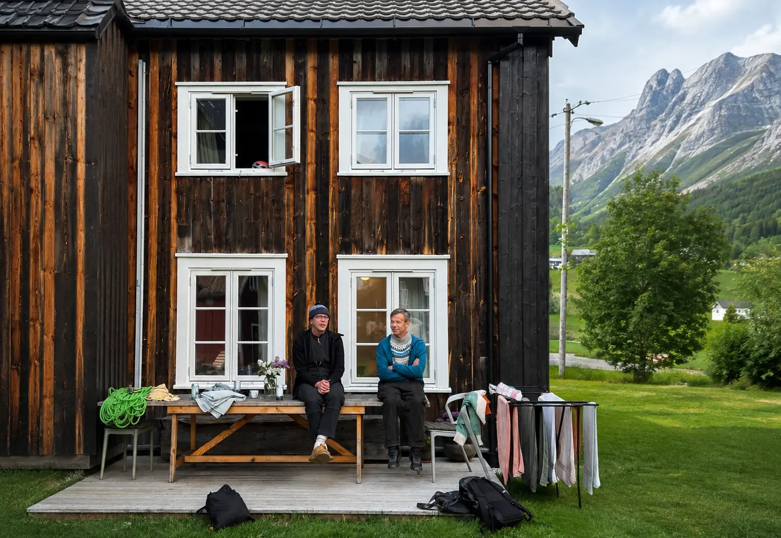 Two people sitting on a wooden bench outside a dark wooden house with white-framed windows, with mountains and greenery in the background.