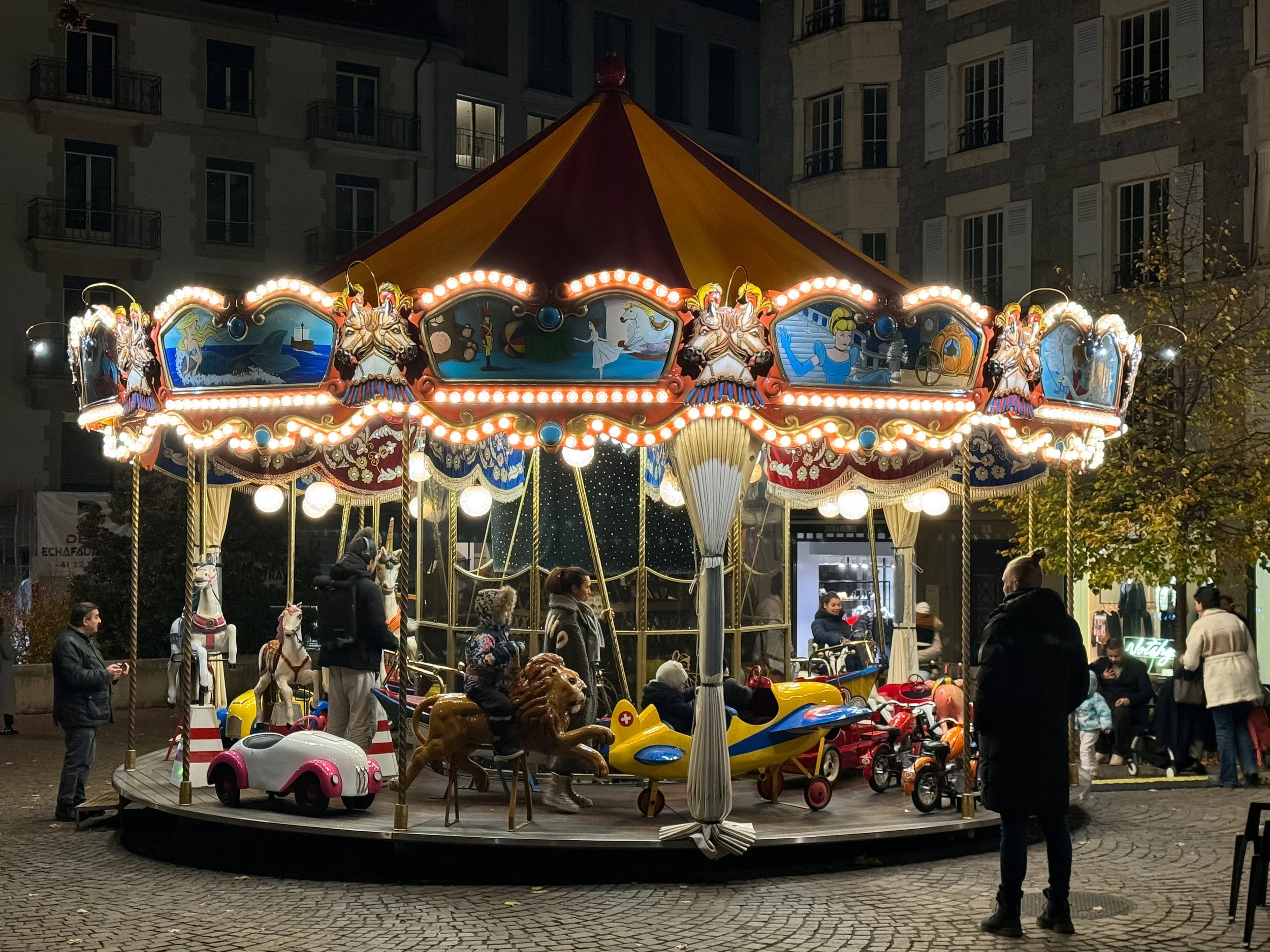 A merry-go-round at a fairground illuminated with bright lights, featuring painted panels with whimsical scenes, and various ride animals including a lion and horses. People are gathered around it, some riding small cars and children on the ride.