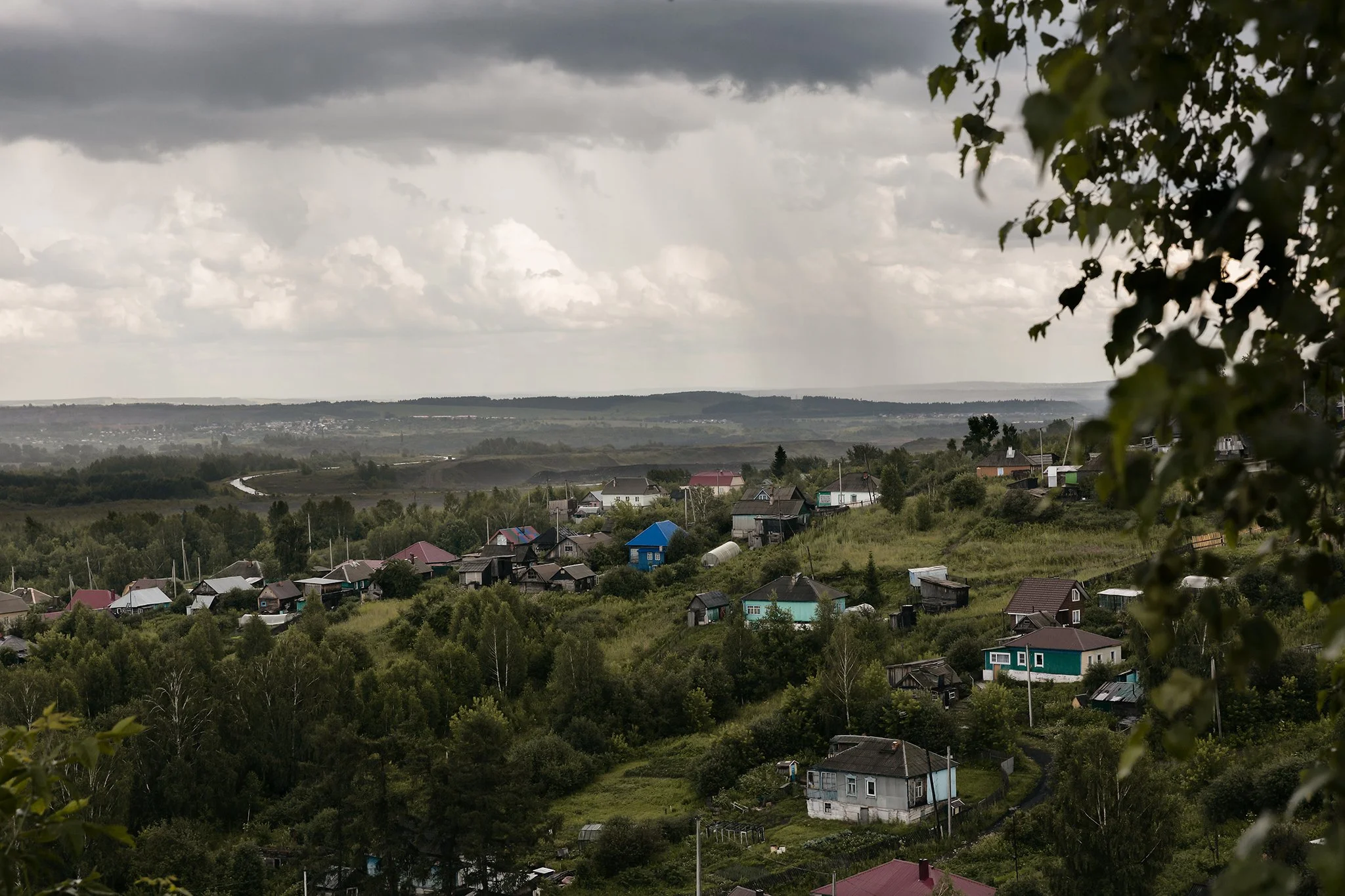 A rural landscape with a cloudy sky, houses scattered on a hillside, and a winding road in the distance. Tree branches and leaves frame the right side of the image.