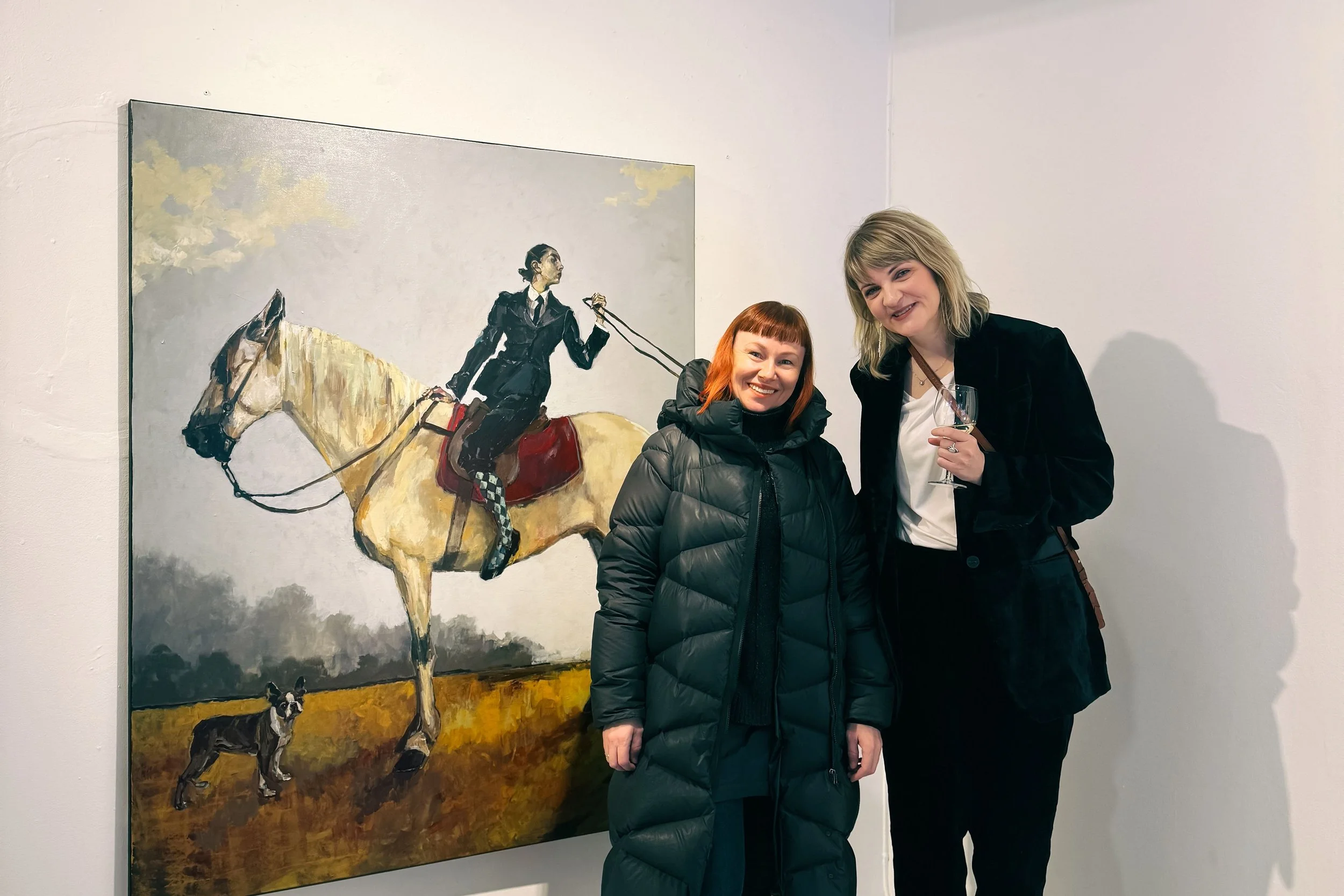 Two women standing in front of a large painting of a woman riding a horse and a dog at a gallery or art exhibition, smiling at the camera.