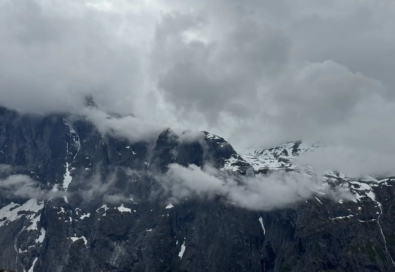 Cloud-covered mountain range with snow patches on dark rocky peaks, under an overcast sky.