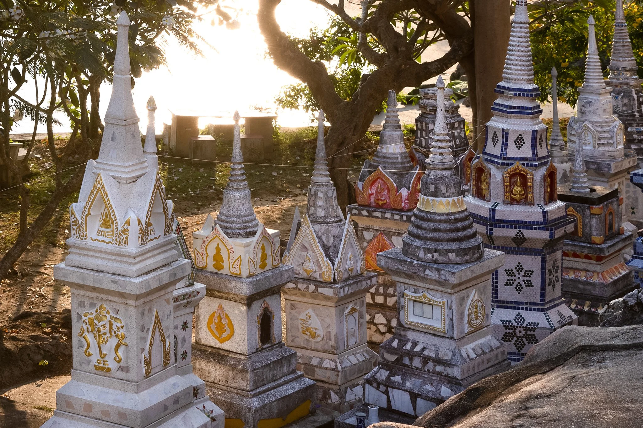 A row of intricately decorated white and colored miniature temple structures with spires, positioned outdoors near trees with sunlight in the background.
