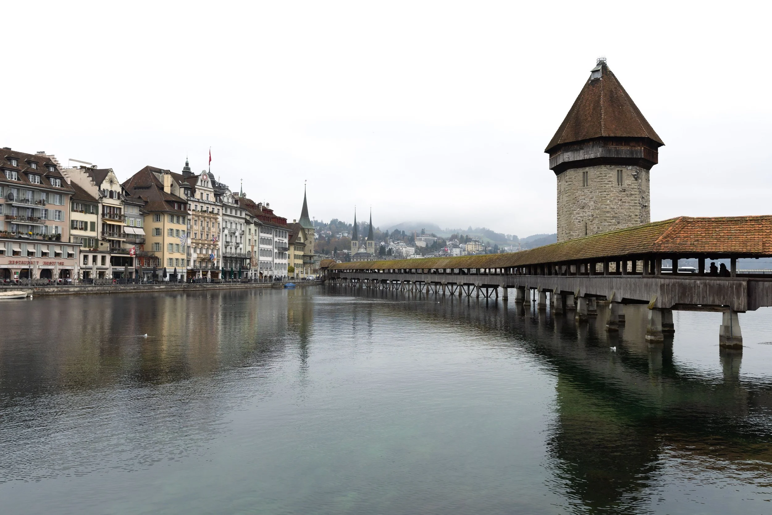 A scenic view of a historic city with colorful buildings, church steeples, and a river with a covered wooden bridge and a medieval stone tower, under a cloudy sky.