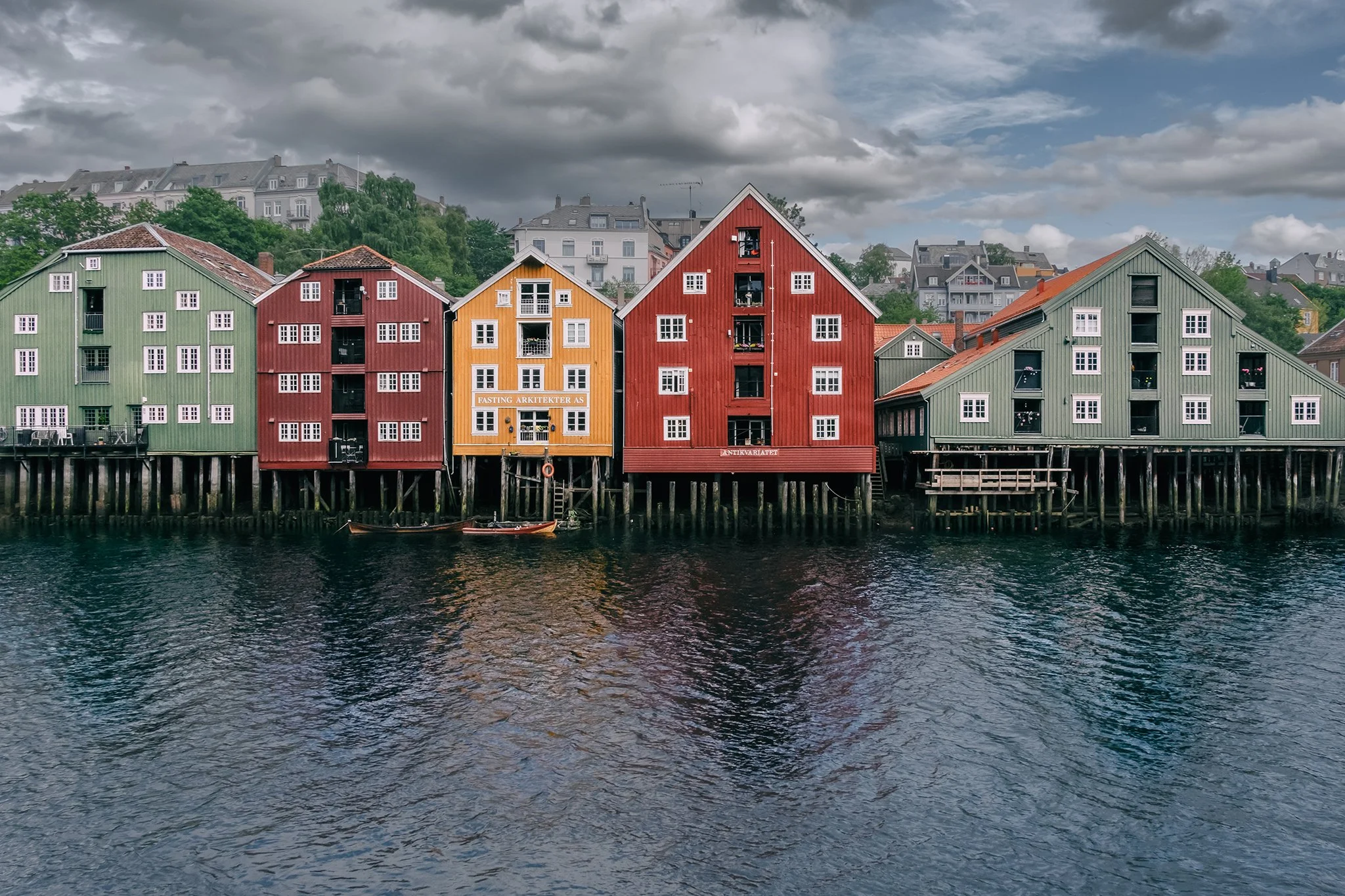 Colorful wooden houses on stilts along a waterfront with water in the foreground and multi-story buildings on a hill in the background under a cloudy sky.