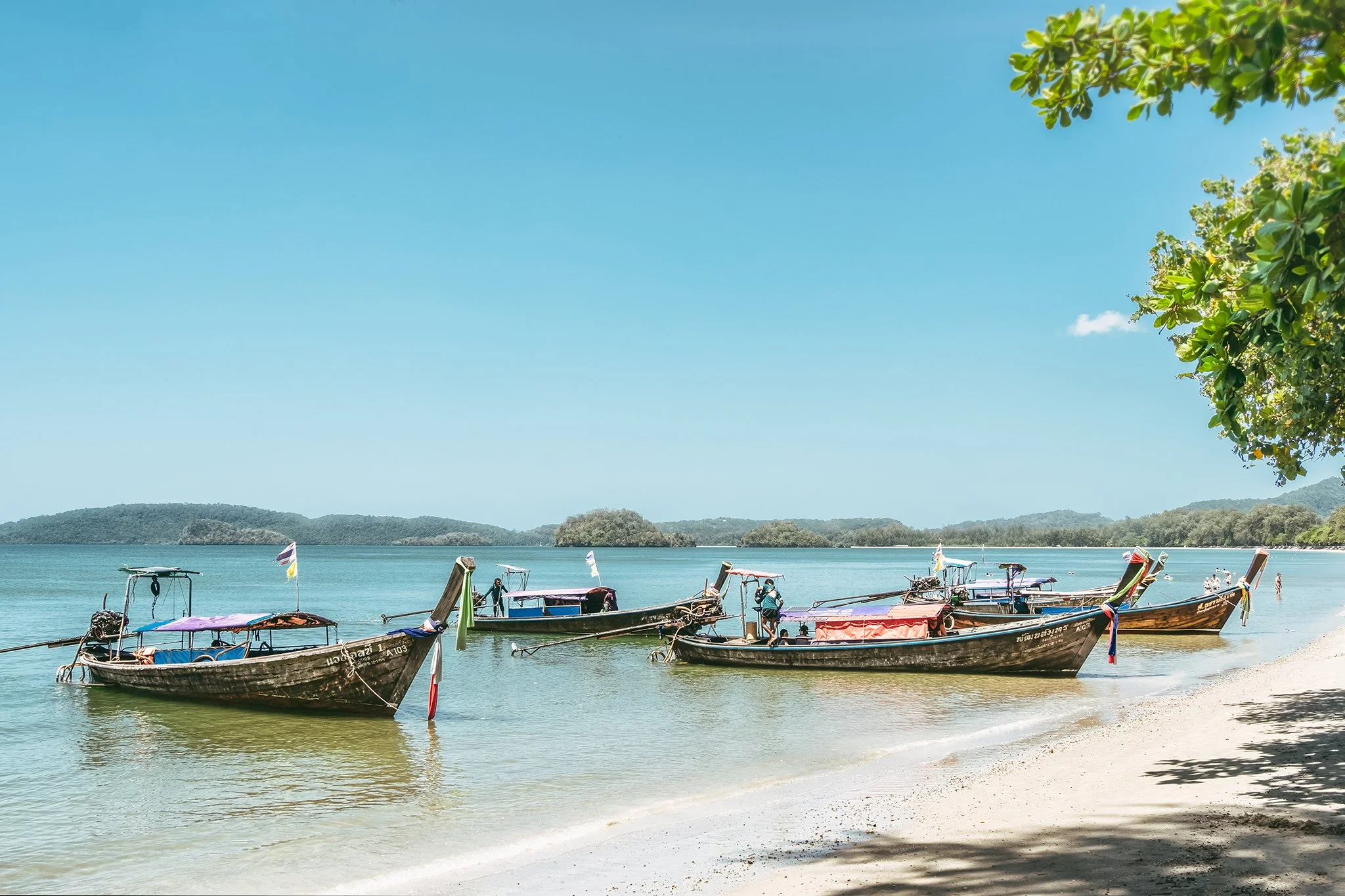 Boats docked at a sandy shore on a calm body of water, with a backdrop of green hills and a clear blue sky with a few clouds.