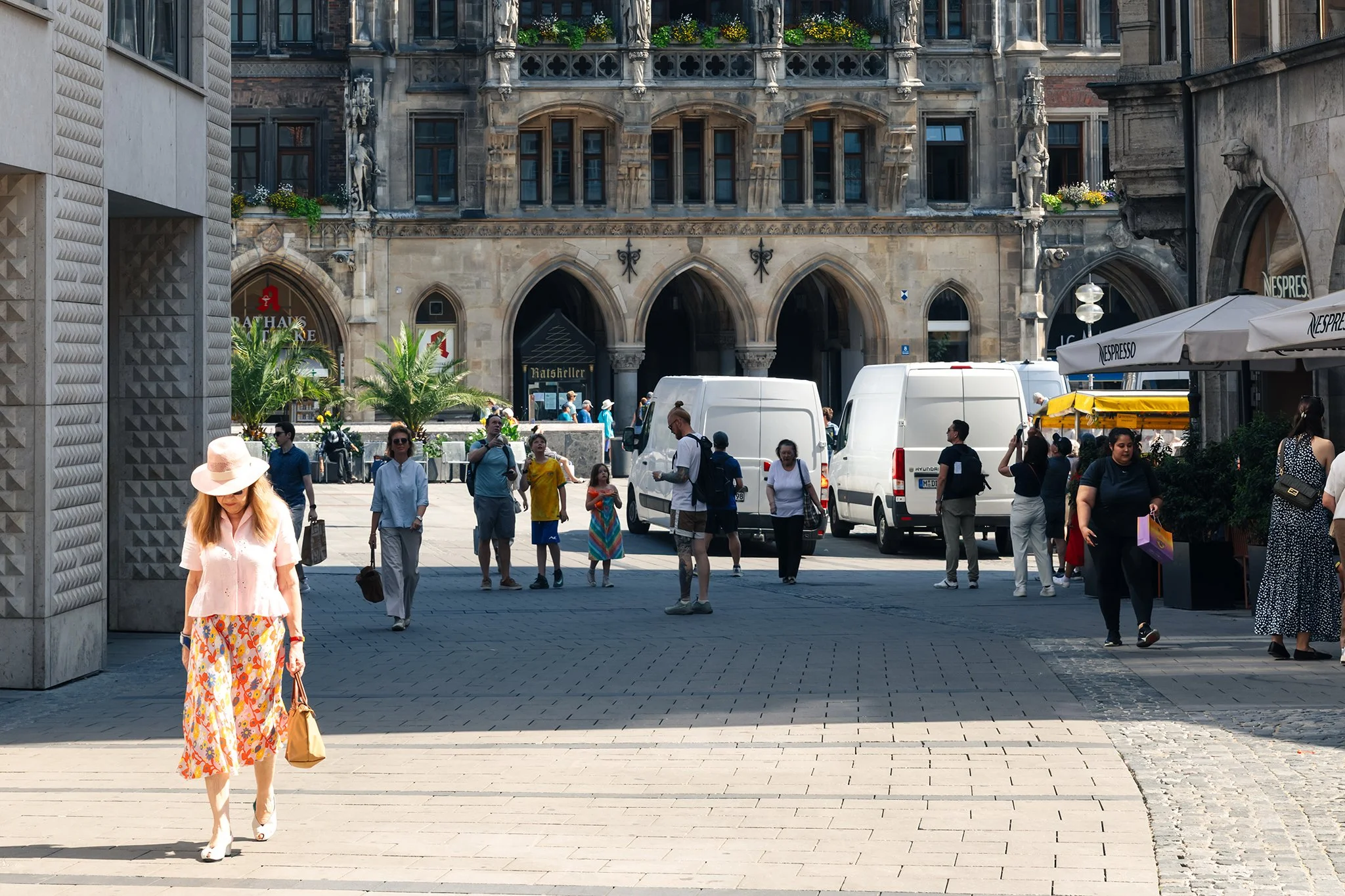 A busy city square with people walking, some carrying shopping bags, and various vehicles parked or passing through. The background features historic architecture with ornate stonework and large windows, along with modern storefronts and outdoor seating areas.