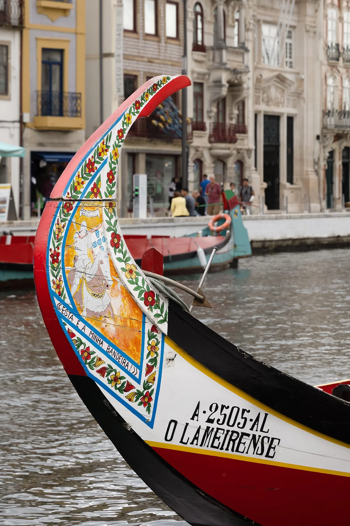 Decorative boat with colorful floral patterns and a crest, docked on a river with historic buildings and people in the background.