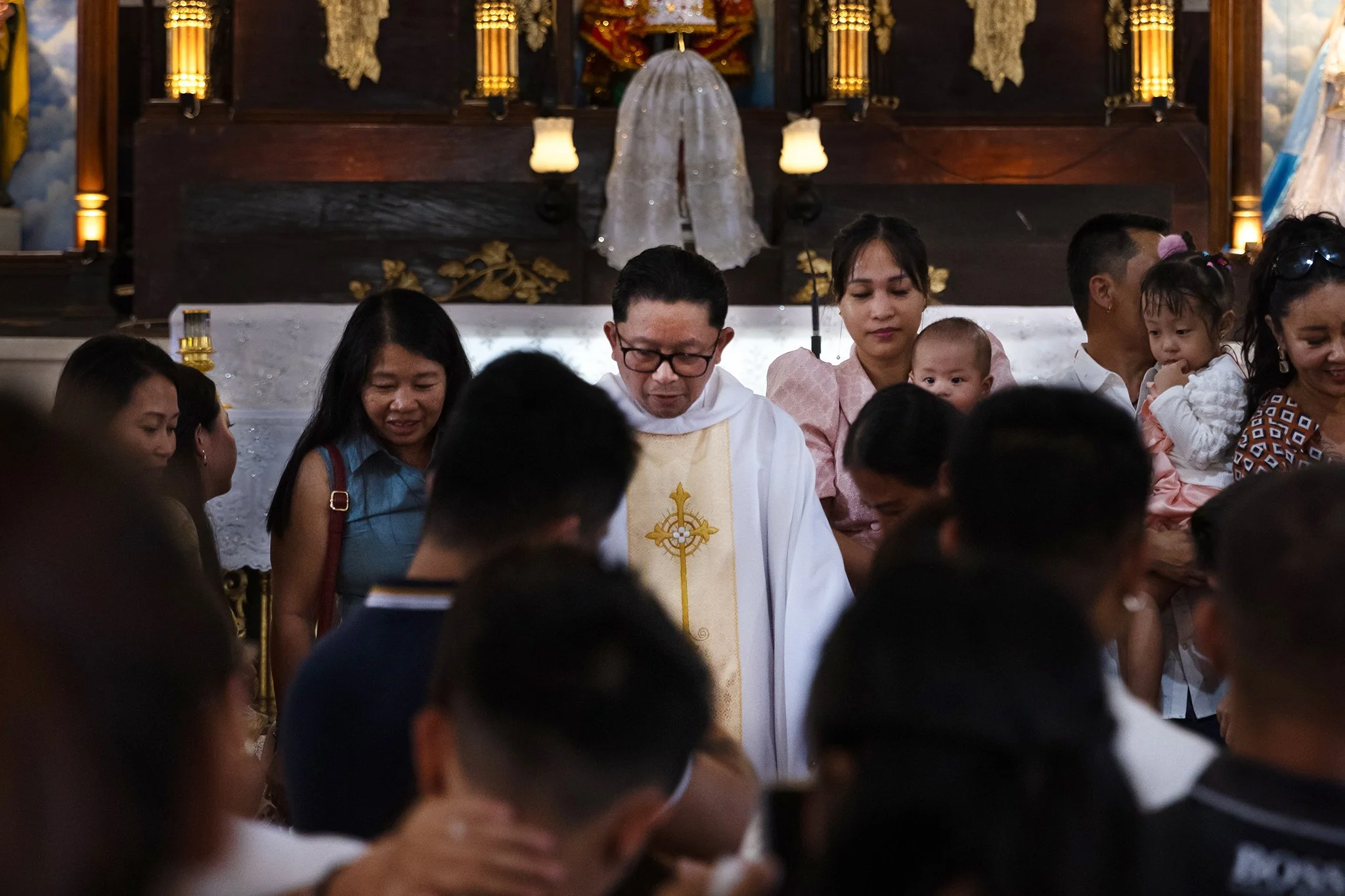 People gathered in a church during a religious ceremony, with a priest in white robes in the center and children and adults surrounding him.