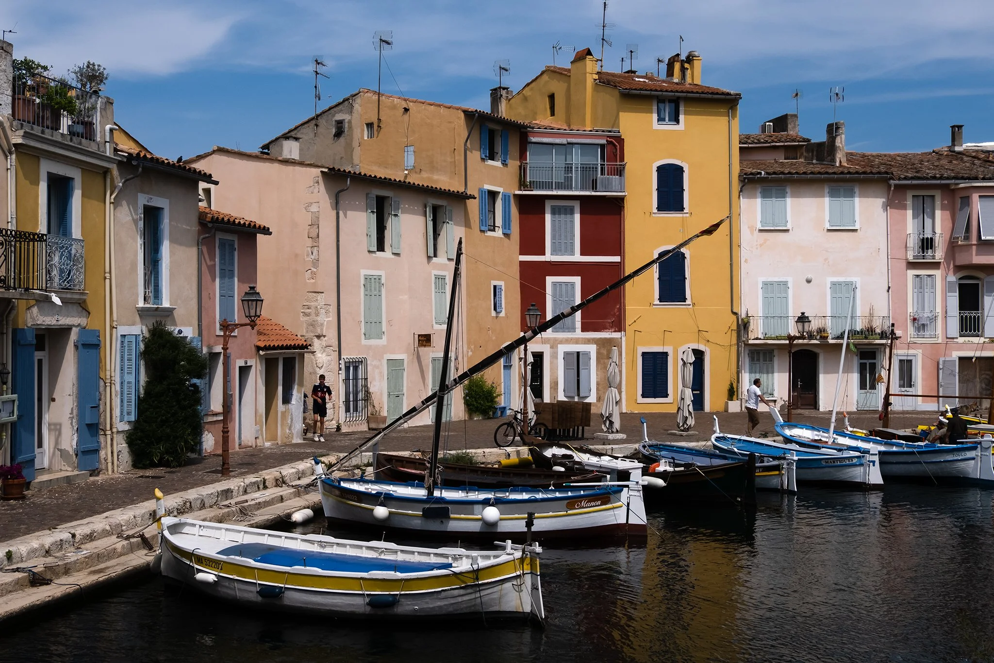 Colorful buildings with blue shutters lining a marina with small boats docked in the water, under a partly cloudy sky.