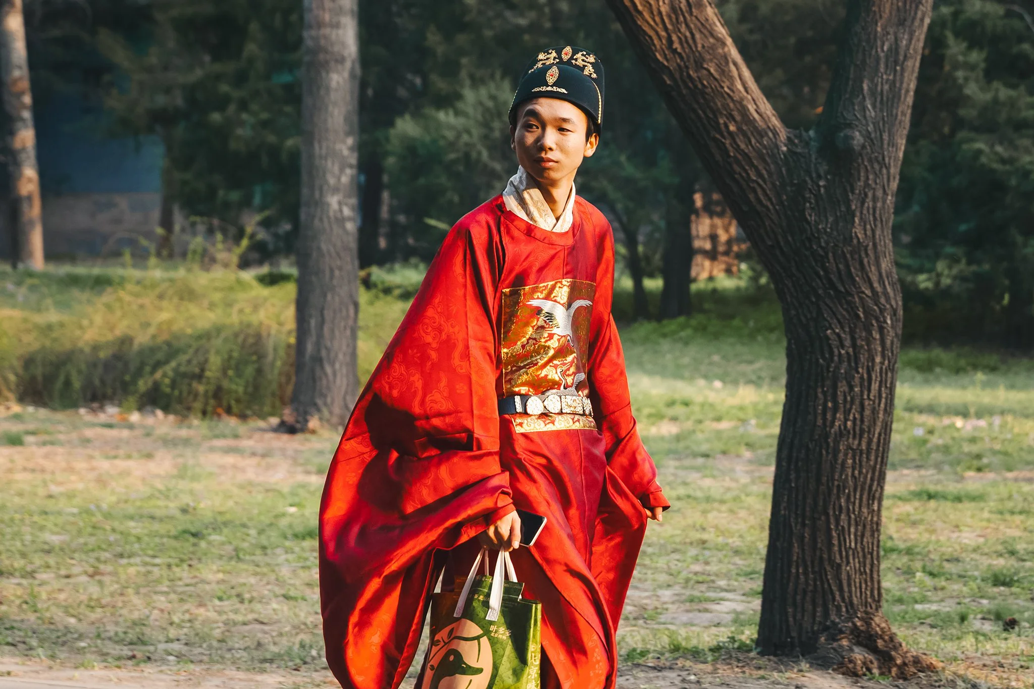 A young person wearing traditional Asian attire, including a black hat with gold embellishments, a red robe with gold and white designs, and a wide belt, walking outdoors in a park with trees. They are carrying shopping bags.