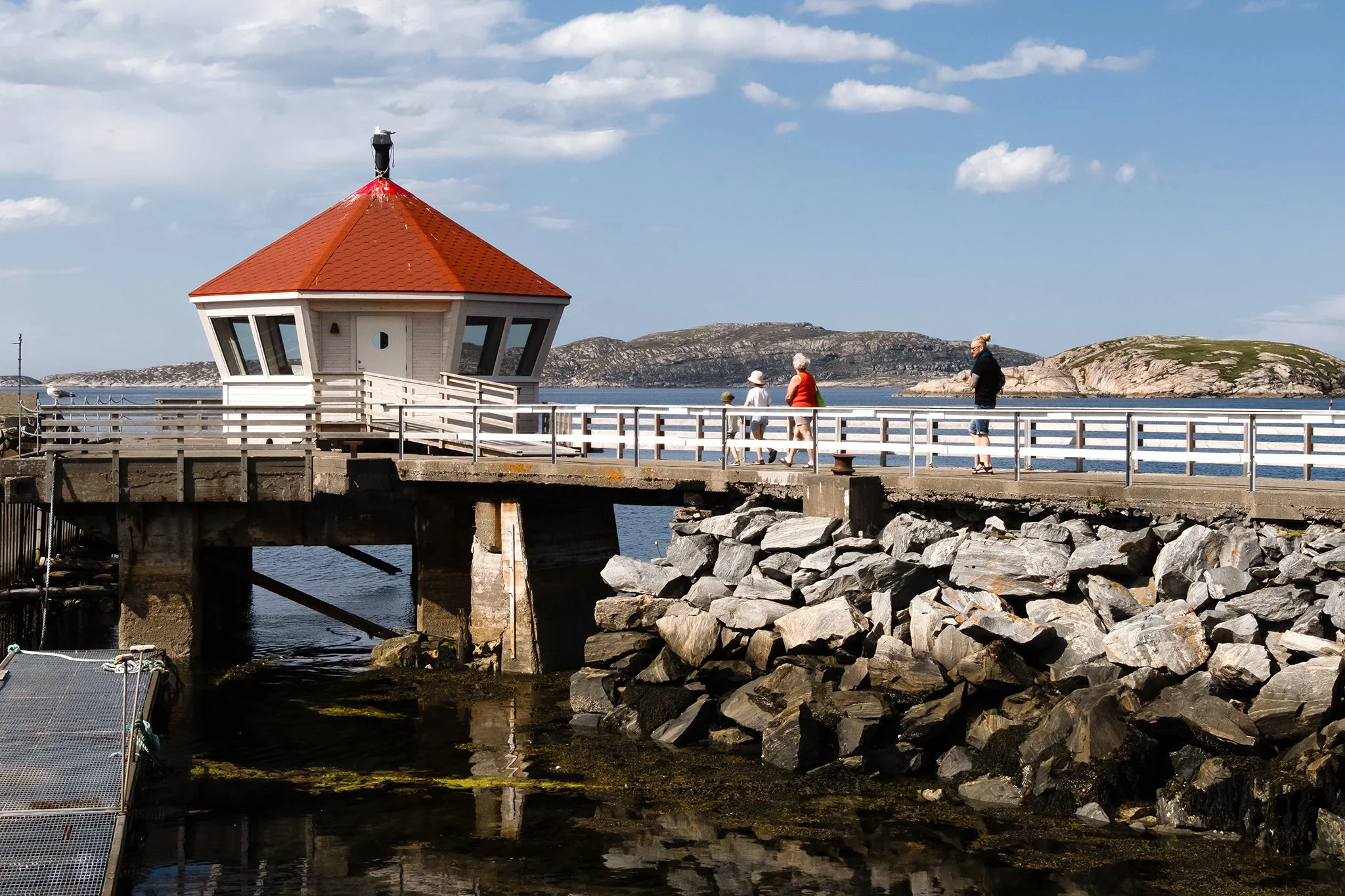 A pier with a small white building with a red roof, overlooking rocky shoreline and water, with three people walking and rocky hills in the background.