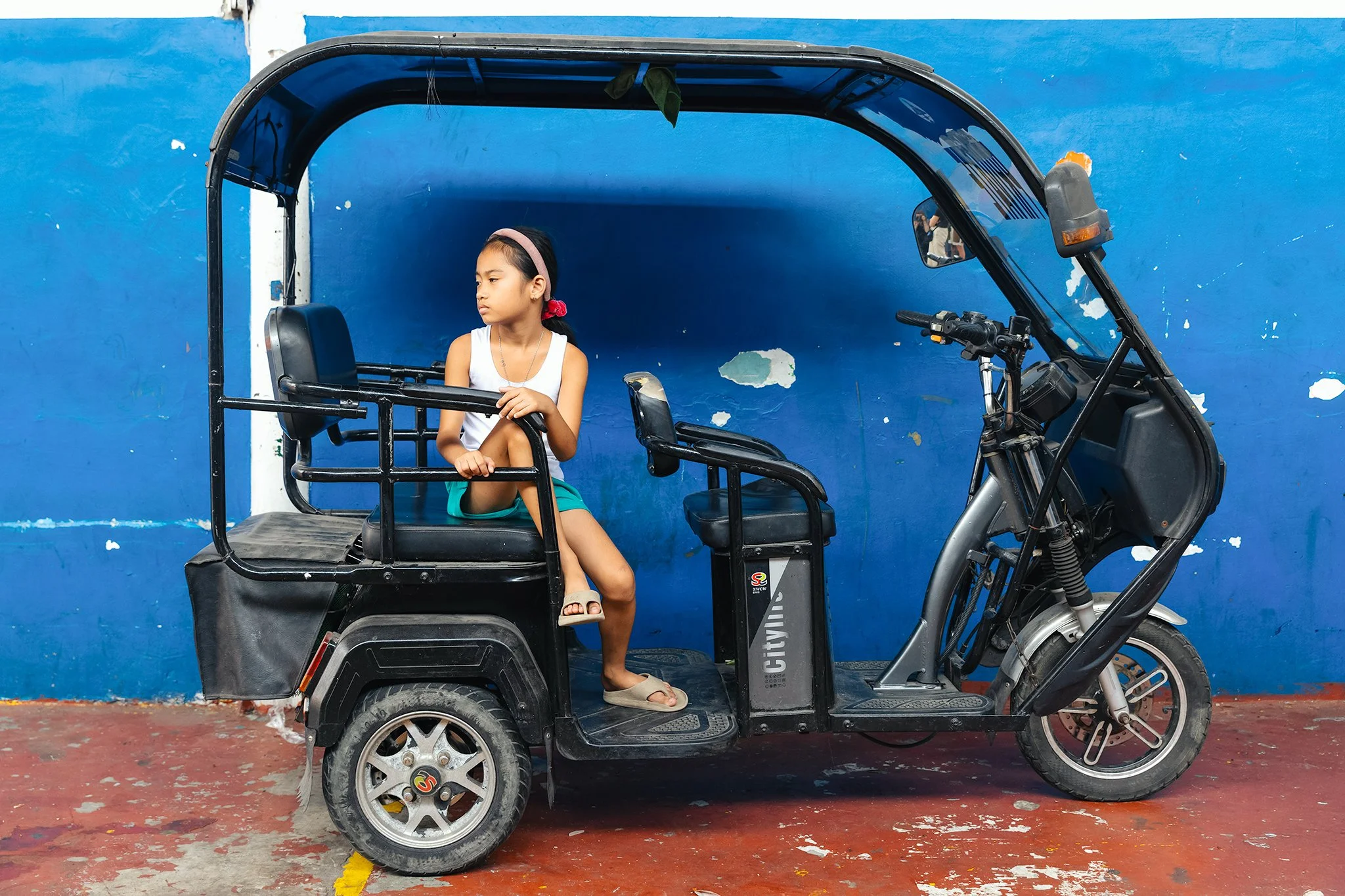 A young girl sitting in the passenger seat of a three-wheeled motorized vehicle, parked against a vibrant blue wall with chipped paint. She wears a white tank top, turquoise shorts, slides, and has a pink headband and a red hair tie with her hair in a ponytail. She appears to be looking off to the side.