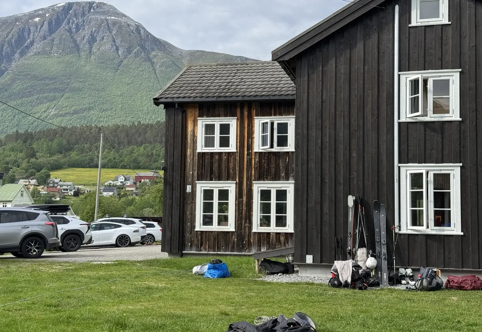 A black wooden house with white-framed windows, surrounded by grass, with mountains and a cloudy sky in the background. Cars are parked nearby, and skis and outdoor gear are leaning against the house.
