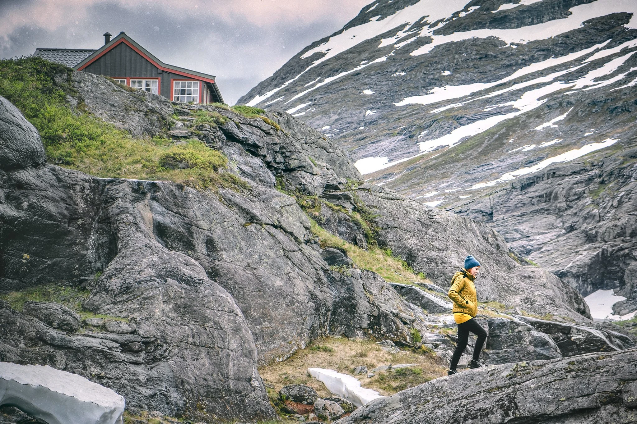 A person dressed in a yellow jacket and black pants walking on rocky terrain in a mountainous area with patches of snow and a house on a cliff.