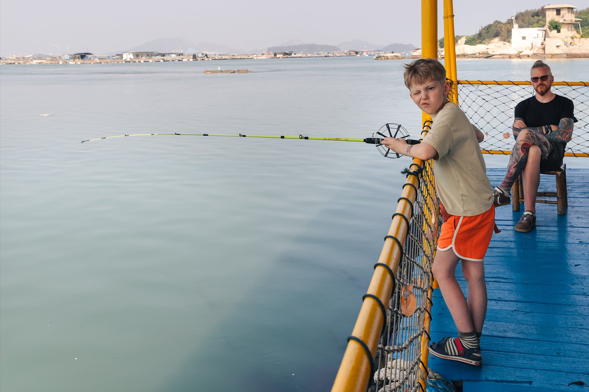 A young boy with brown hair fishing off the edge of a boat, with an older man with tattoos sitting in the background on the boat, overlooking calm water with buildings and mountains in the distance.