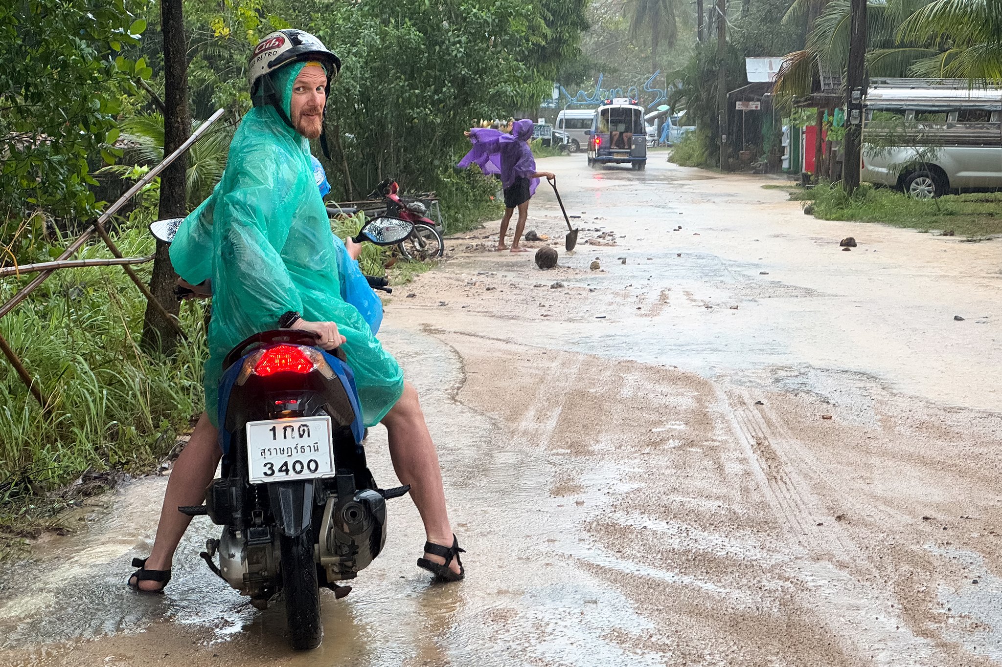 A man in a turquoise raincoat and sandals is sitting on a motorcycle on a muddy, flooded street. He is wearing a helmet and looking back. A woman with a purple umbrella is walking ahead, managing the floodwaters with a stick. Vehicles are visible in the background on a rain-soaked road.