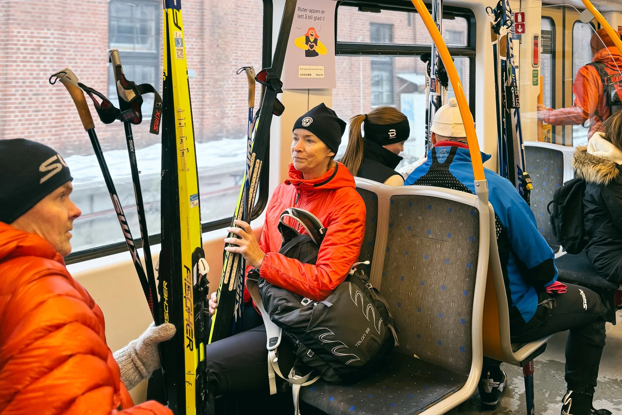 People seated inside a bus holding cross-country skis, with winter clothing and hats, and winter snow visible through the windows.