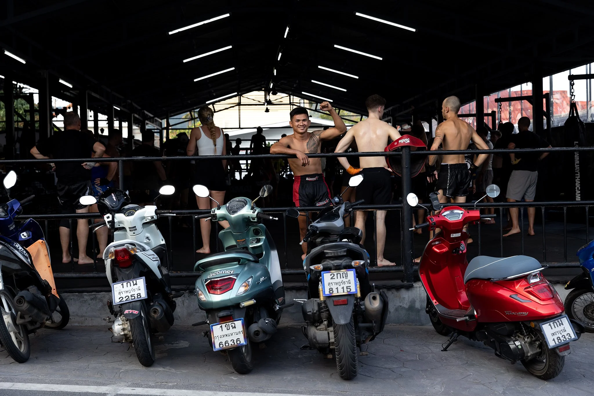 Group of young people, including shirtless men, at a motorcycle repair shop or garage, with scooters parked in front and motorcycles inside.
