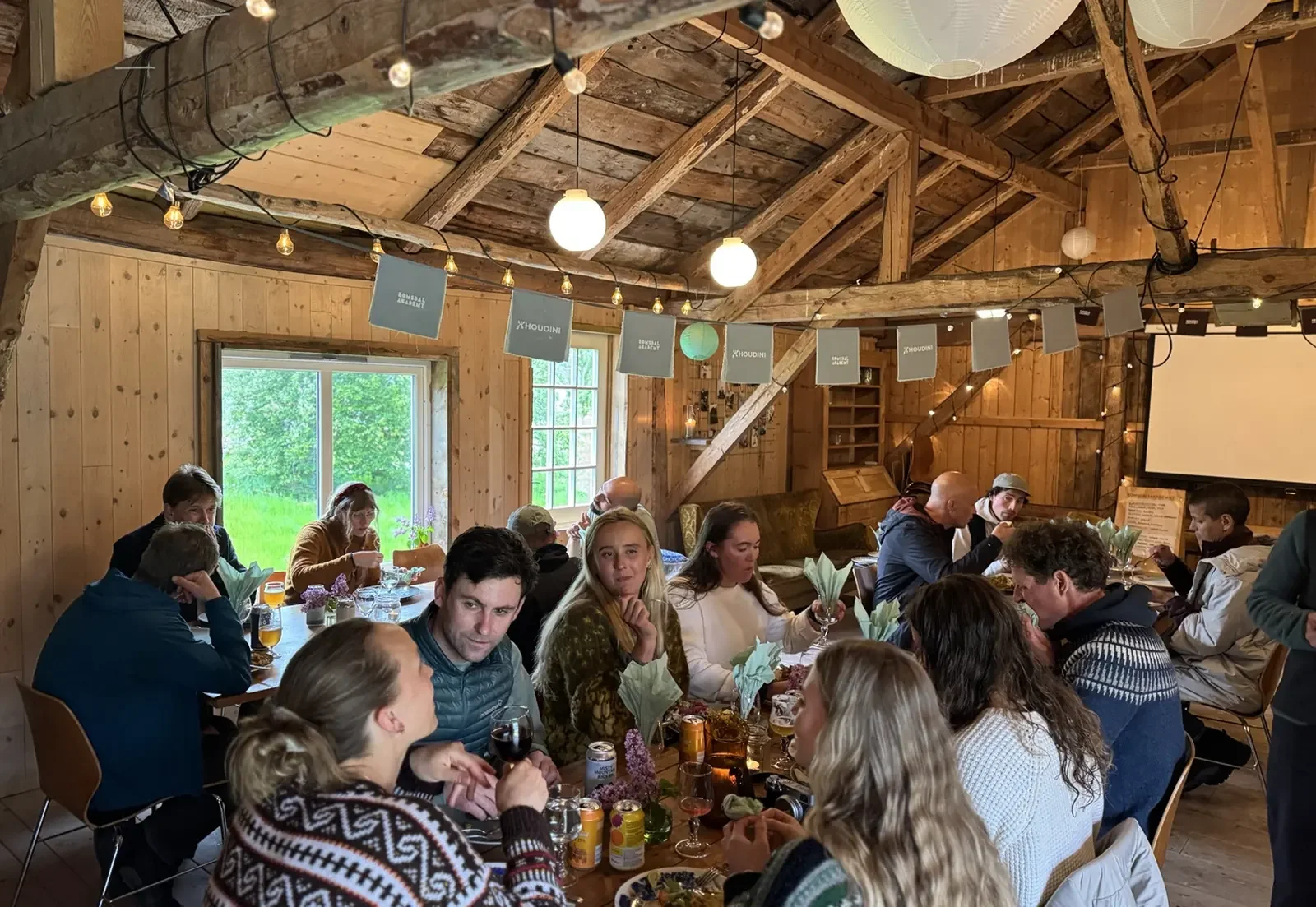 Group of people sitting at a long wooden table inside a rustic wooden lodge during daytime, with some holding drinks and engaged in conversation, with decorative string lights and paper lanterns hanging from the ceiling.