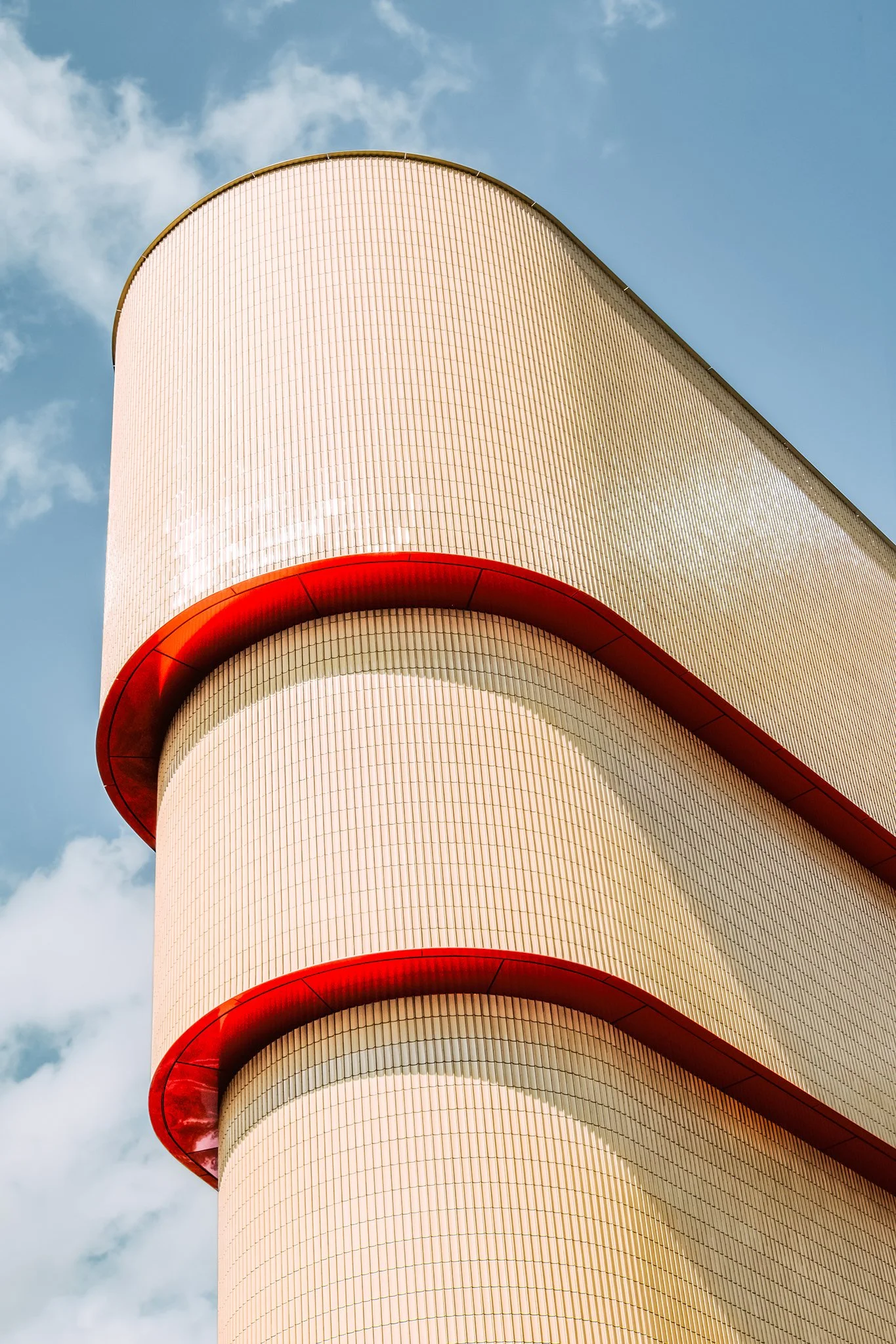 Close-up of a modern building with curved beige tiles and red accents, against a background of blue sky with clouds.