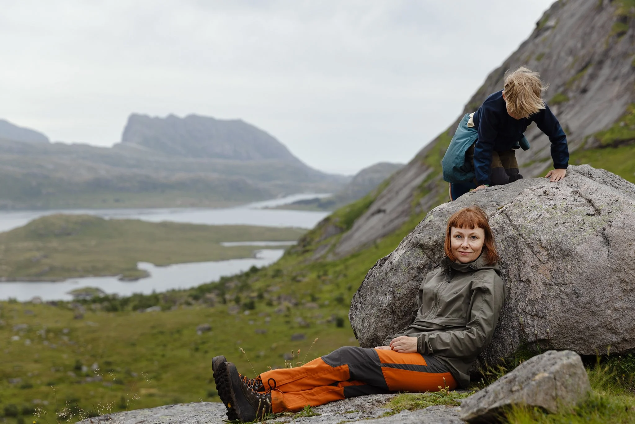 A woman and a young boy outdoors on rocks, with a scenic landscape of mountains, water, and grassy terrain in the background.