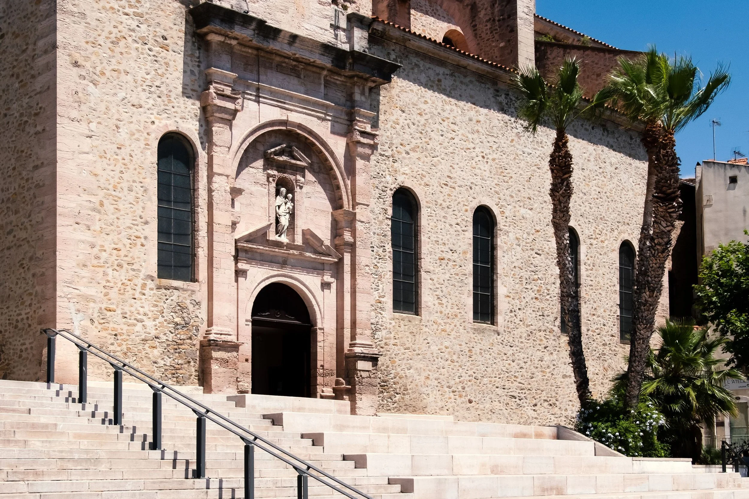 The image depicts the facade of a historic stone church with arched windows, a sculptural niche with a religious statue, and a staircase leading up to the entrance, flanked by palm trees and a clear blue sky in the background.