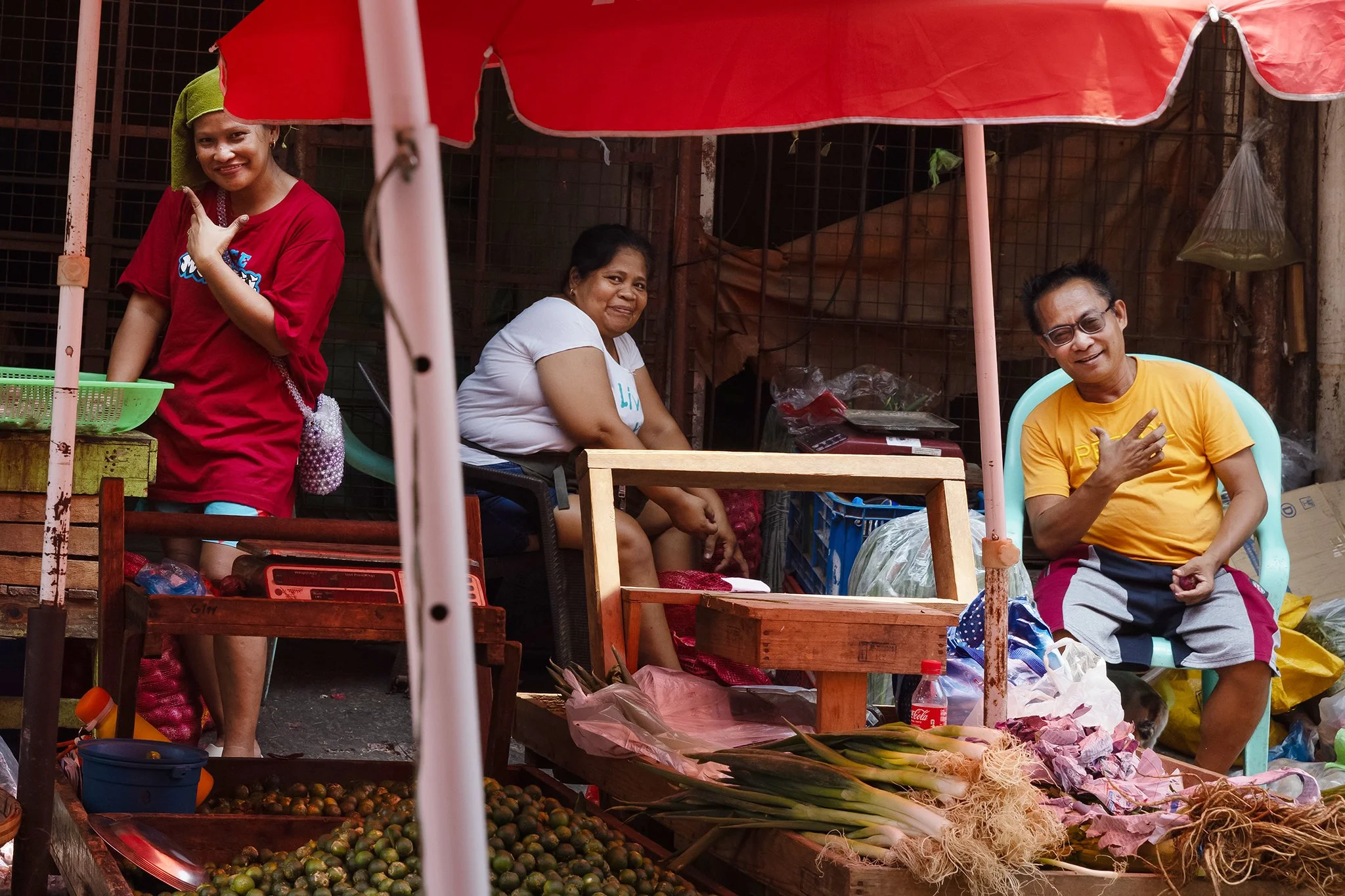 Three women at a market stall, one standing and two sitting, with various fruits and vegetables displayed in front of them under a red canopy.