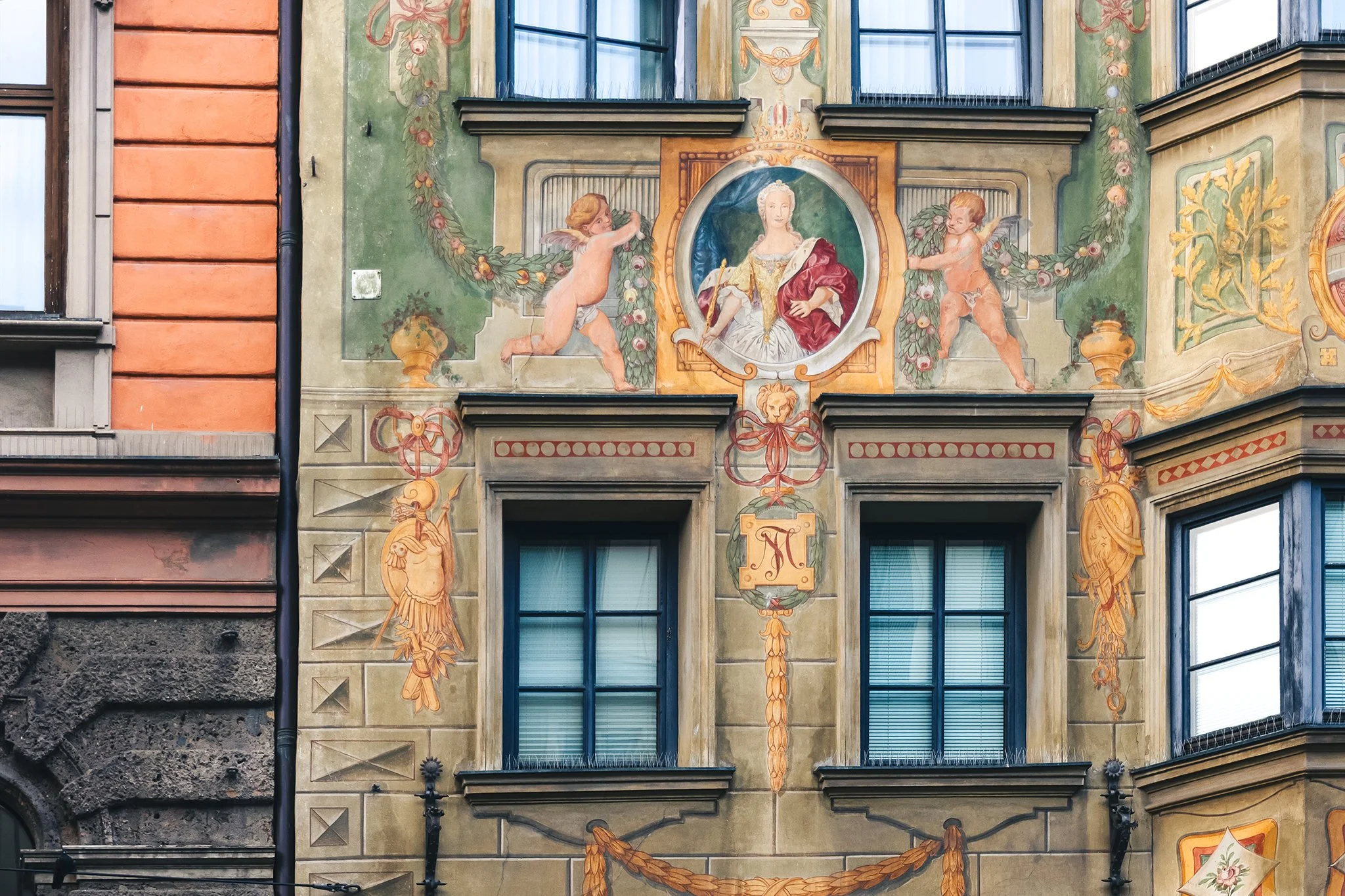 Decorative mural painted on a building's exterior wall featuring a noblewoman in a circular frame, flanked by two cherubs holding floral garlands, with ornate designs and draperies surrounding the scene.
