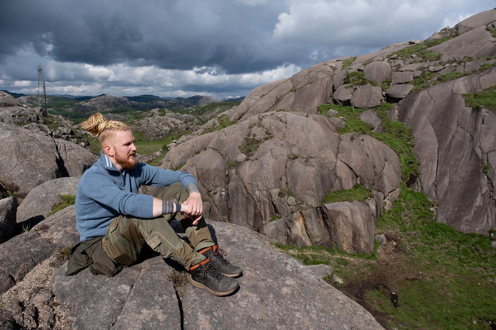 A man with blond hair and a beard sitting on rocks in a mountainous landscape with large boulders, green patches, and a cloudy sky.