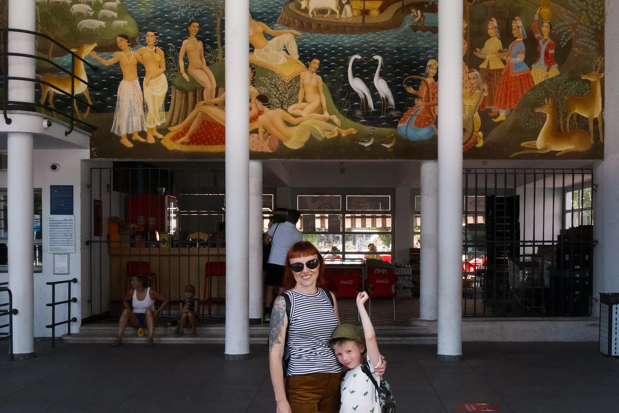 A woman with tattoos on her arms and sunglasses is smiling and holding the hand of a young boy with a backpack. They are standing in front of a large mural that depicts a scenic outdoor scene with women, animals, and water, with some people sitting and standing in front of the mural inside a building.