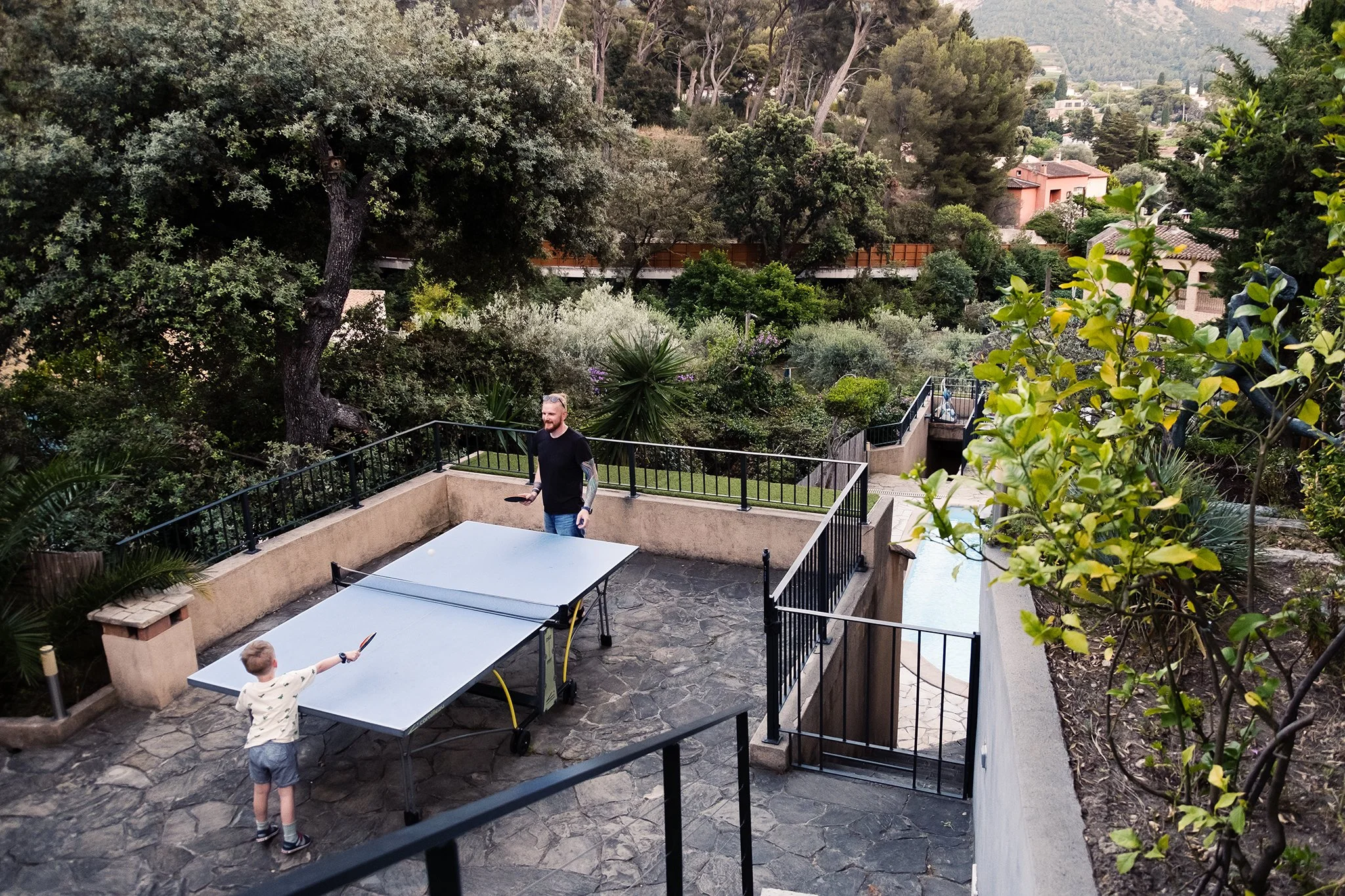 A man and a child playing table tennis on a patio with lush green trees and bushes in the background.