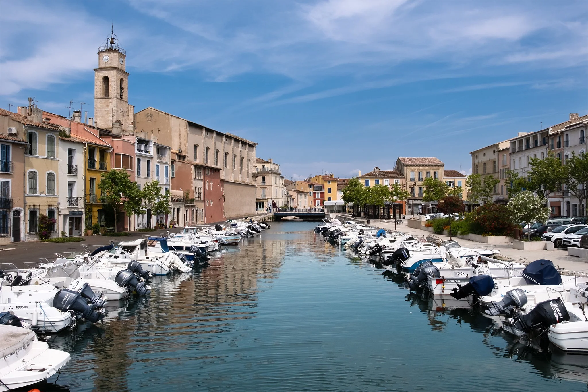 A marina with boats docked in calm water, colorful buildings lining the waterfront, and a clock tower against a blue sky with wispy clouds.